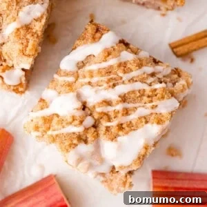A square piece of rhubarb cake with icing drizzle on top, surrounded by pieces of rhubarb and a cinnamon stick on a white surface.