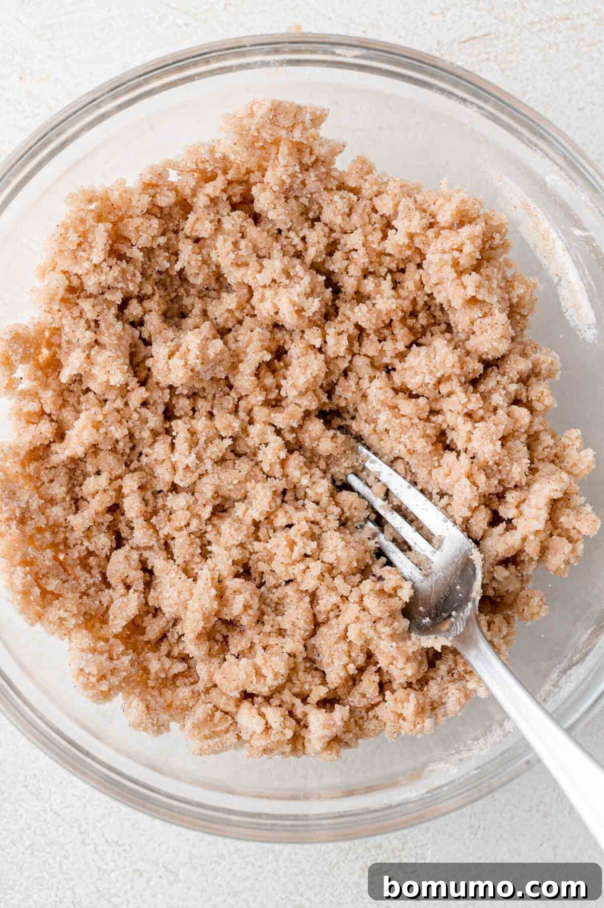 Tangy Rhubarb Cake 6 A transparent glass bowl filled with a crumbly mixture of brown sugar, butter, and cinnamon, with a metal fork resting in the mixture, ready for use as a streusel topping.