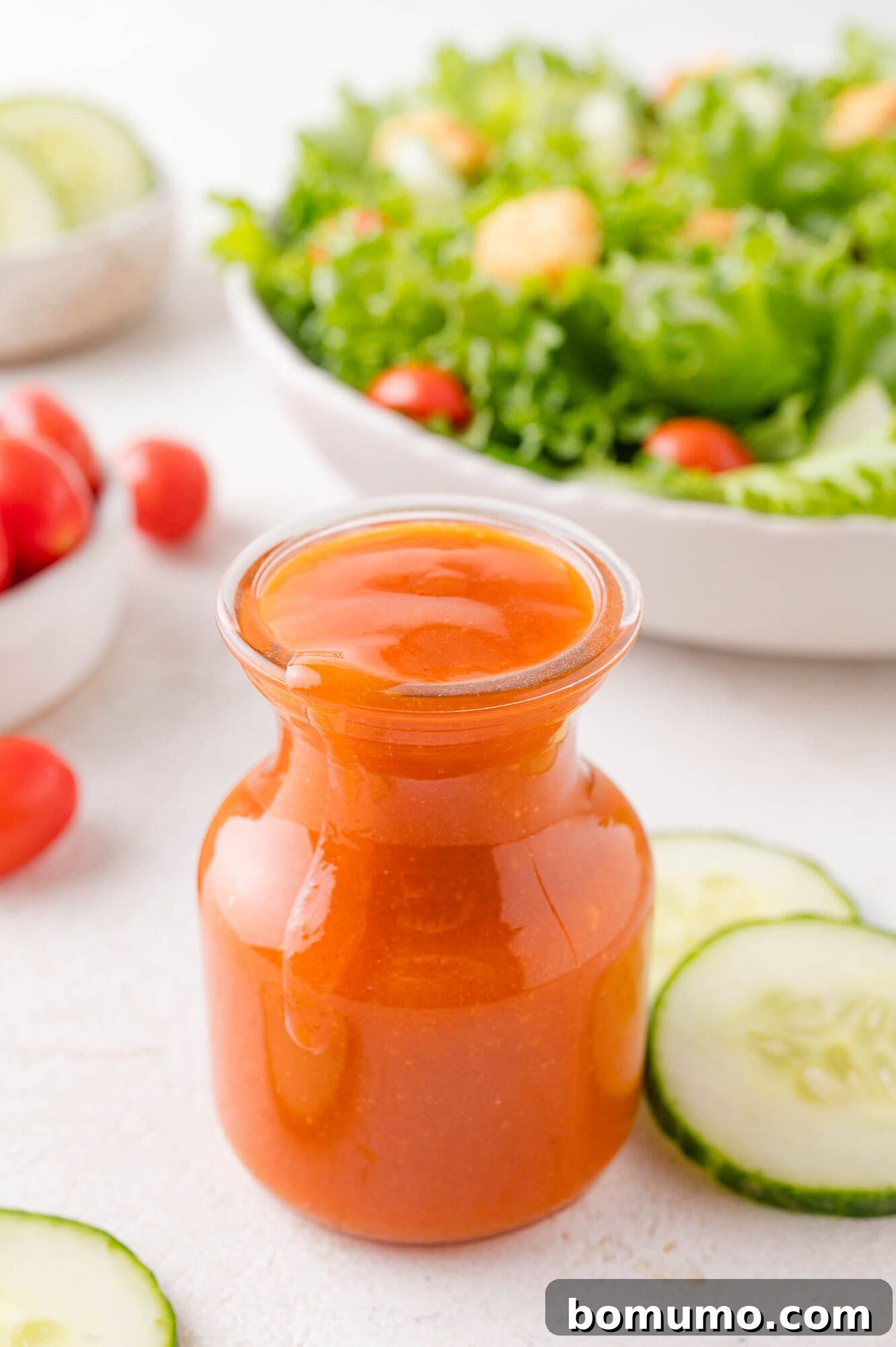 Sunny Tang Dressing 3 A glass jar filled with Calalina salad dressing sits on a white surface, with cucumber slices and a bowl of green salad in the background.
