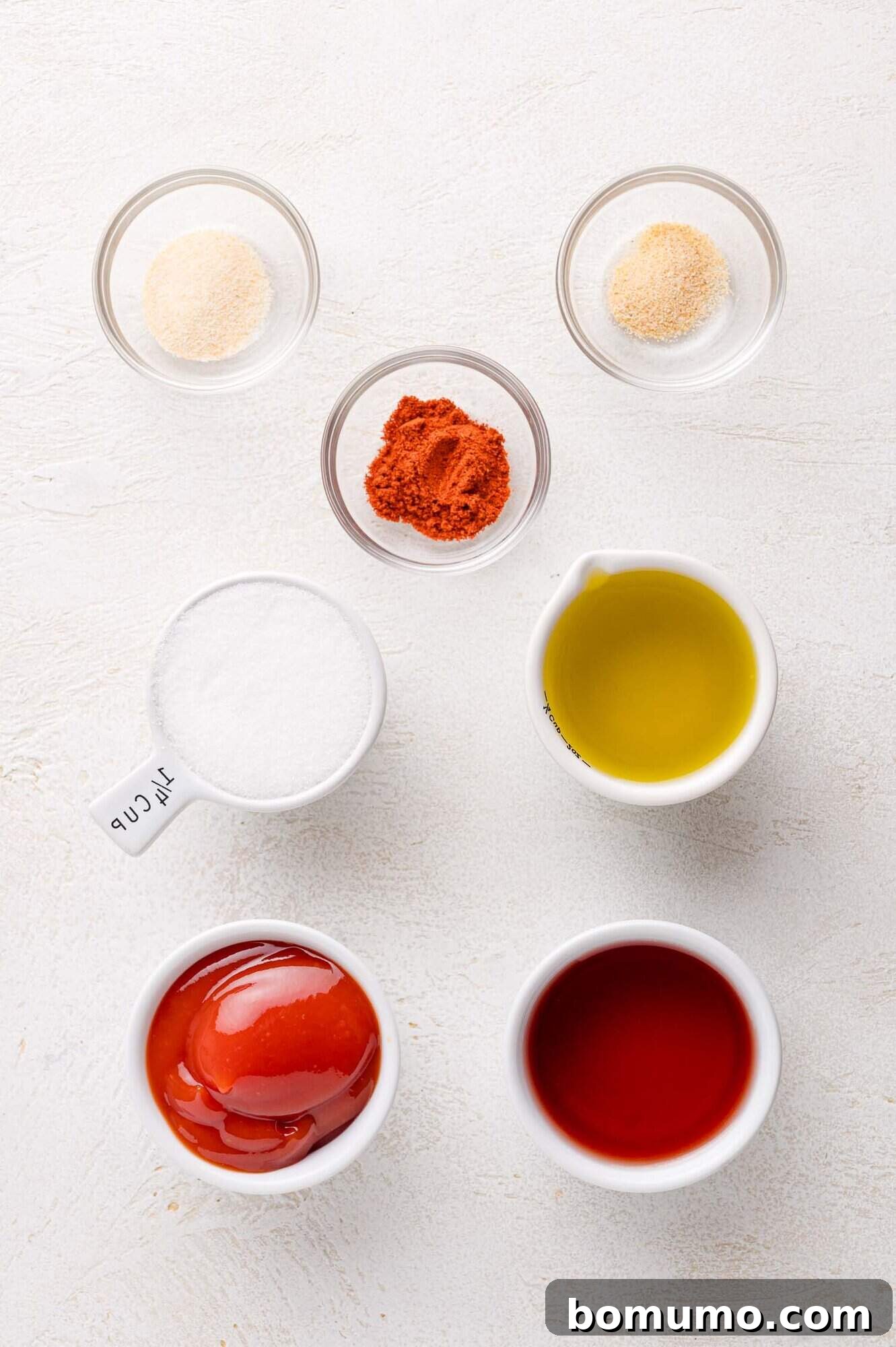 Sunny Tang Dressing 4 Seven small bowls on a white surface contain garlic powder, onion powder, paprika, sugar, olive oil, ketchup, and vinegar, arranged in a neat pattern.