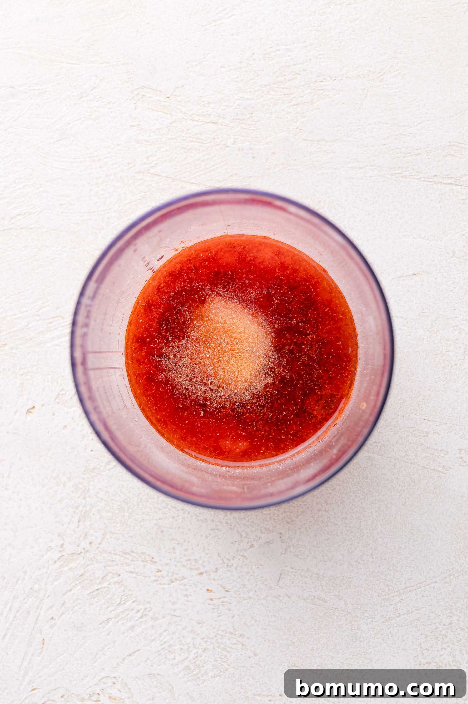 Sunny Tang Dressing 5 A clear bowl containing a red liquid mixture with a mound of granulated seasoning in the center, viewed from above on a light textured surface.
