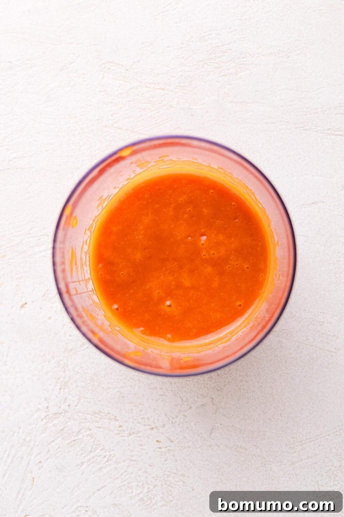 Sunny Tang Dressing 6 A clear glass bowl filled with smooth, bright orange salad dressing on a white textured surface, viewed from above.