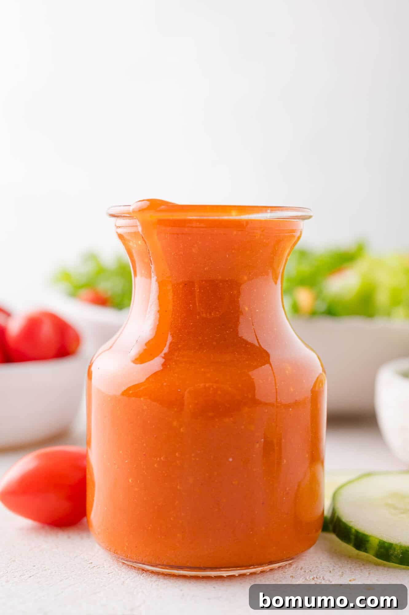 Sunny Tang Dressing 7 A glass jar filled with orange colored salad dressing sits on a white surface, with vegetables like tomatoes and cucumber in the background.