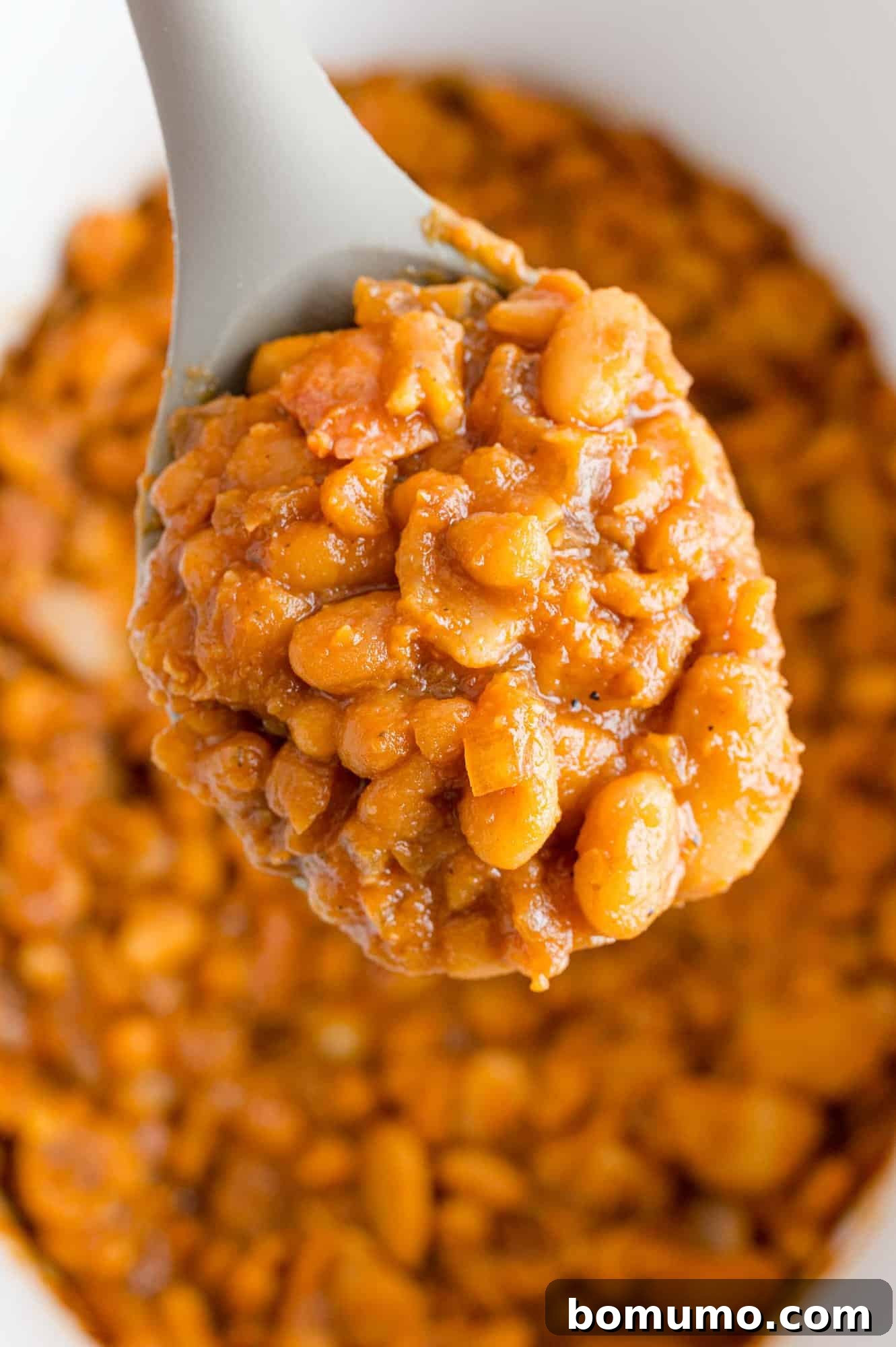 A spoonful of rich, saucy crock pot baked beans held over a larger bowl, showcasing their inviting texture and color.