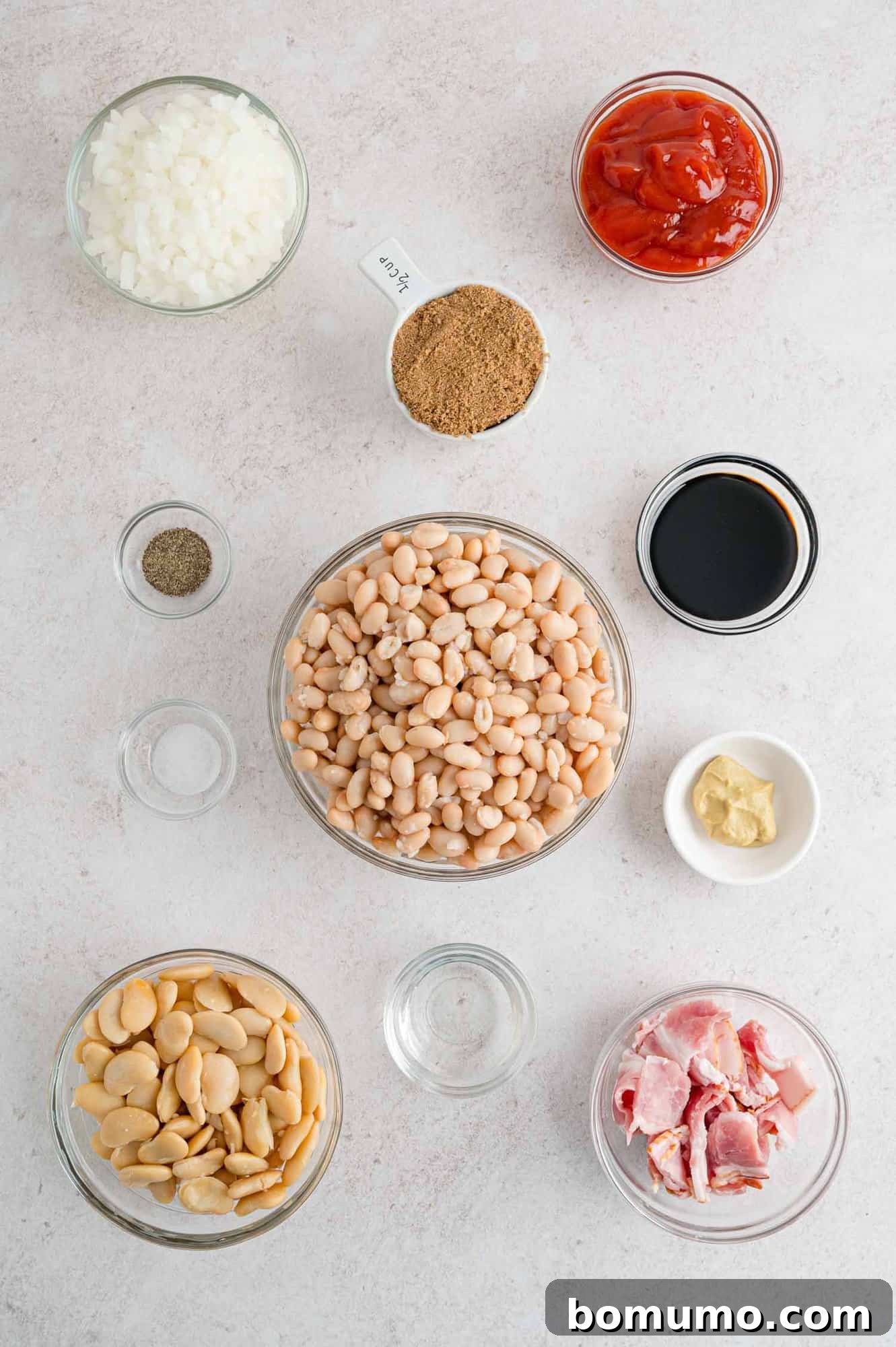 Overhead shot displaying an array of raw ingredients for baked beans: canned beans, bacon, onion, ketchup, molasses, and brown sugar.
