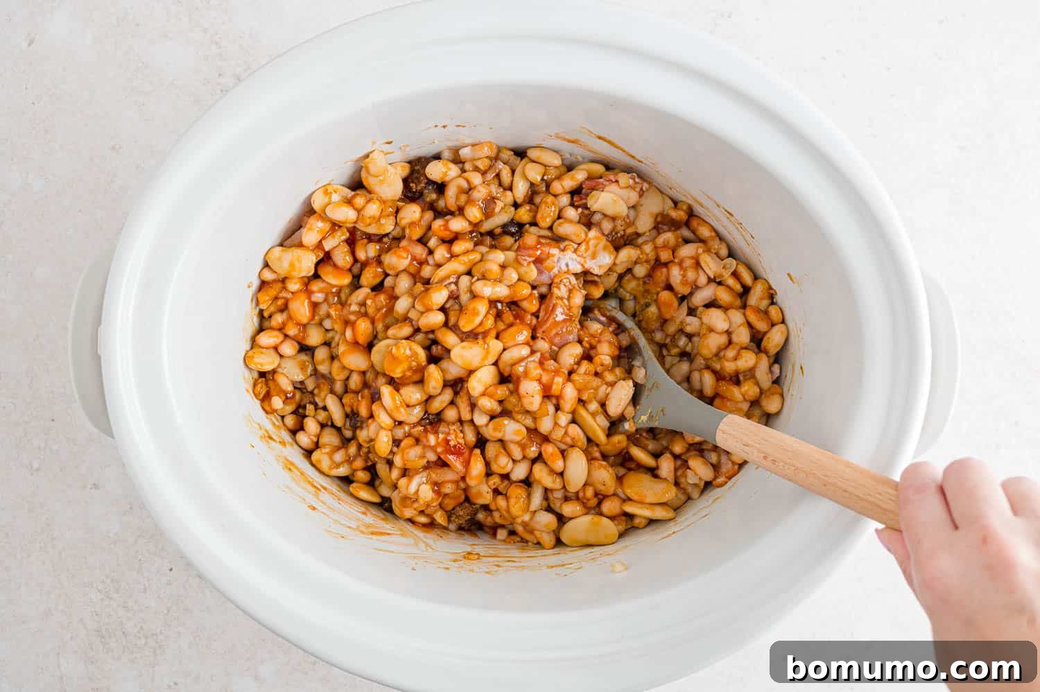 Hands stirring the combined ingredients for baked beans in a white slow cooker, ensuring everything is well mixed.