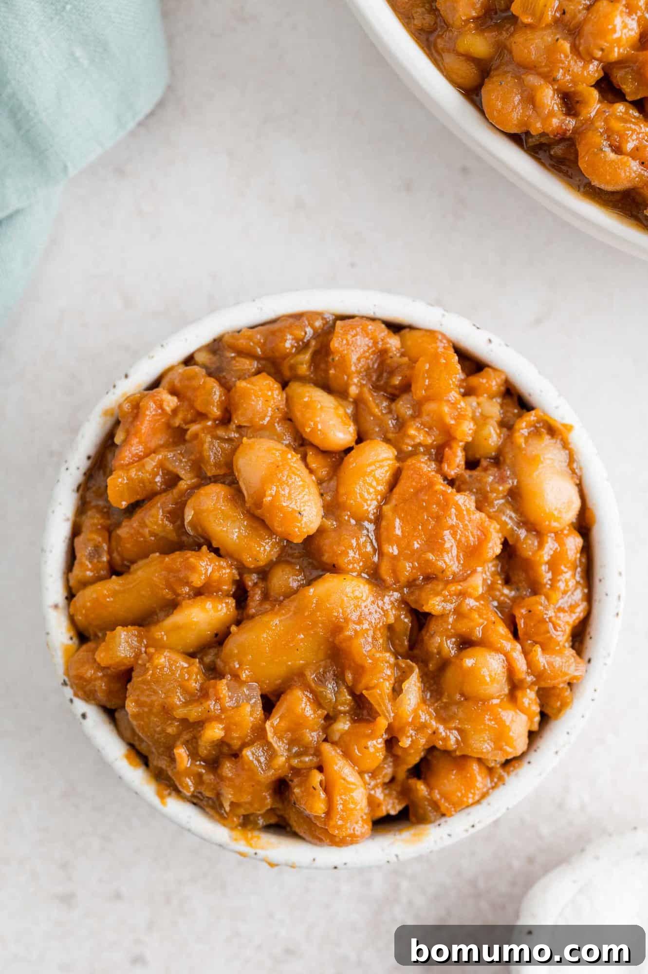 A small white bowl brimming with hearty crock pot baked beans, garnished with a sprinkle of fresh herbs.