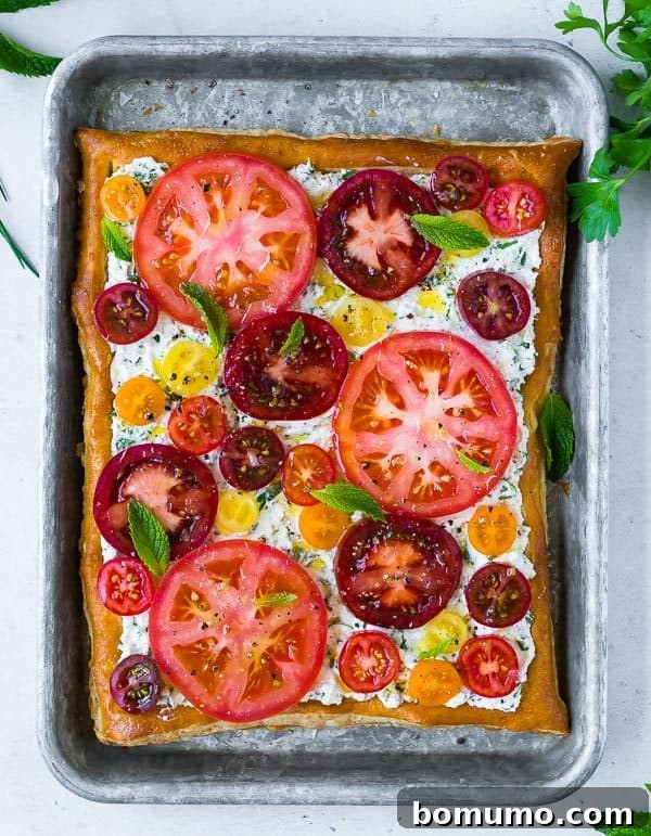 An overhead view of a vibrant fresh tomato tart, showcasing a beautiful arrangement of colorful heirloom tomatoes, fresh mint leaves, and creamy herbed ricotta on a flaky puff pastry. The presentation is inviting and suggests a taste of summer.