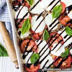 overhead image of caprese quinoa bake, sprinkled with fresh basil and drizzled with balsamic reduction. Wooden spoon and linen next to white baking dish.