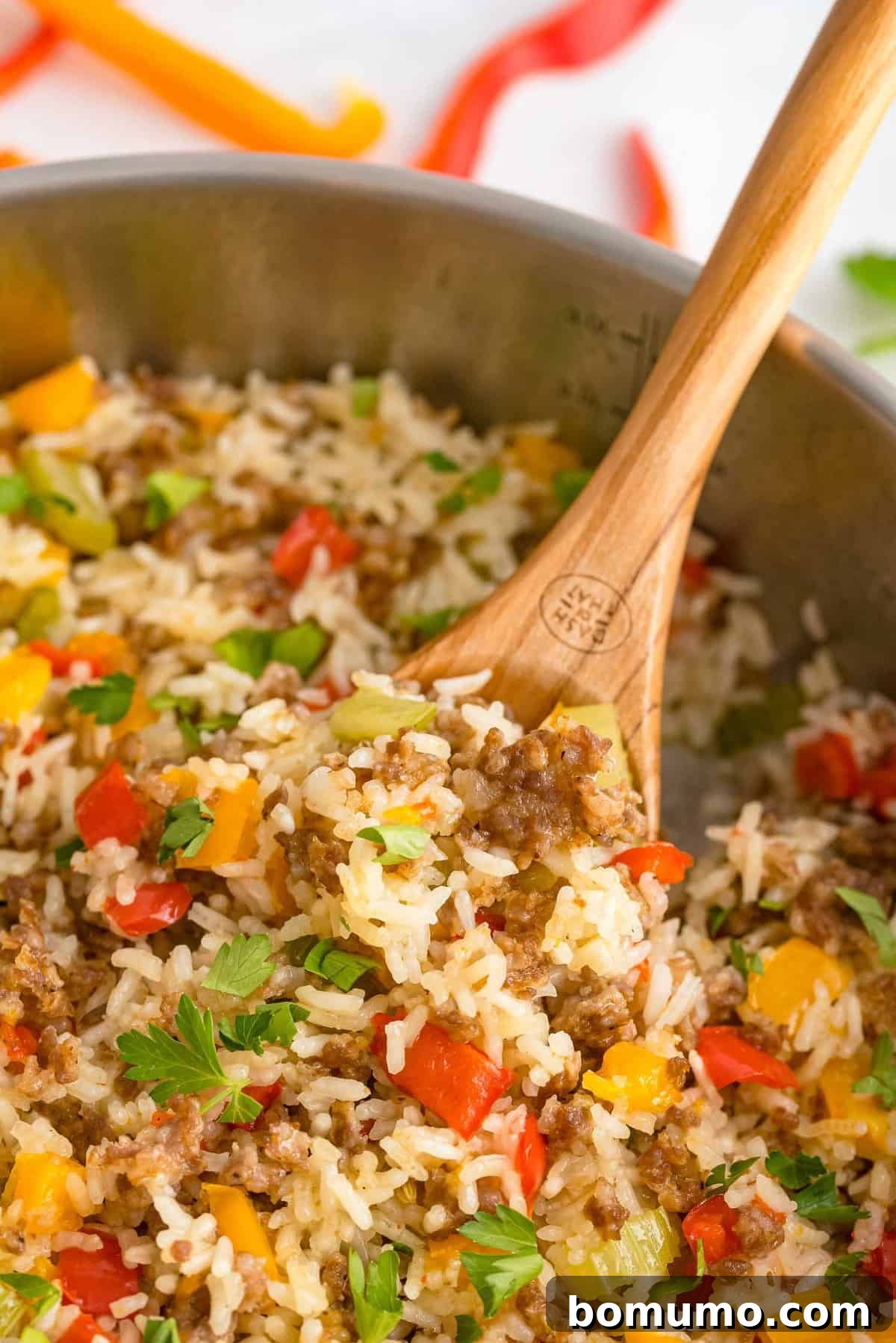 Close up of vibrant rice, savory sausage, and colorful bell peppers in a single skillet, ready to be served with a wooden spoon.