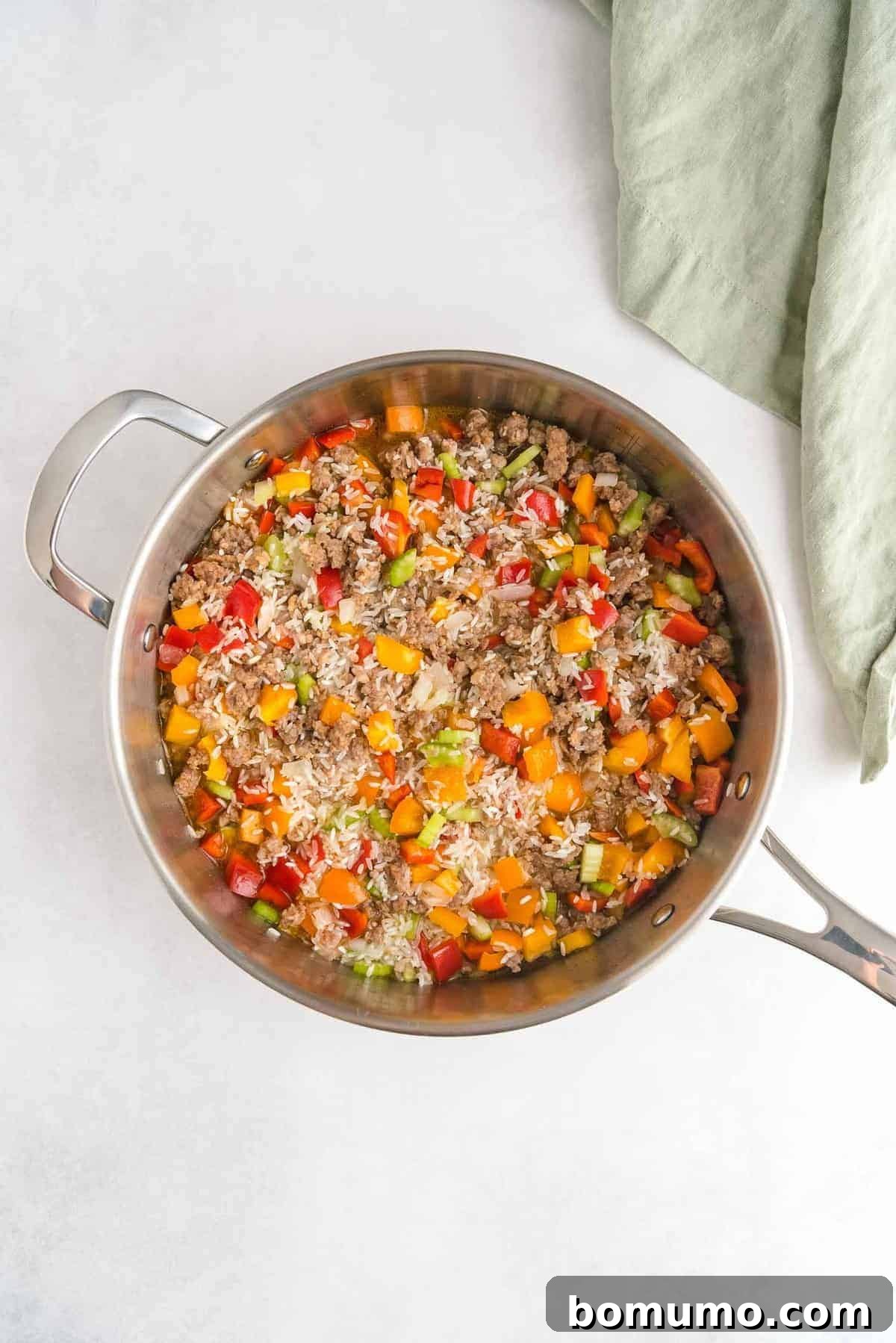 An overhead view of a large sauté pan brimming with colorful bell peppers, savory sausage, and uncooked rice, all simmering together in liquid before covering.