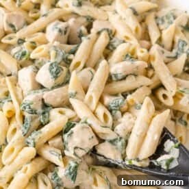 Close-up of creamy penne pasta mixed with pieces of chicken and spinach, being served with a black spoon.