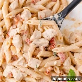 Close-up of creamy penne pasta with pieces of chicken and sun-dried tomatoes, garnished with fresh herbs, served on a black spoon.