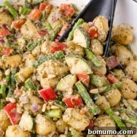 Gnocchi mixed with chopped red bell peppers, asparagus, zucchini, red onion, pine nuts, and pesto sauce, in a pan.
