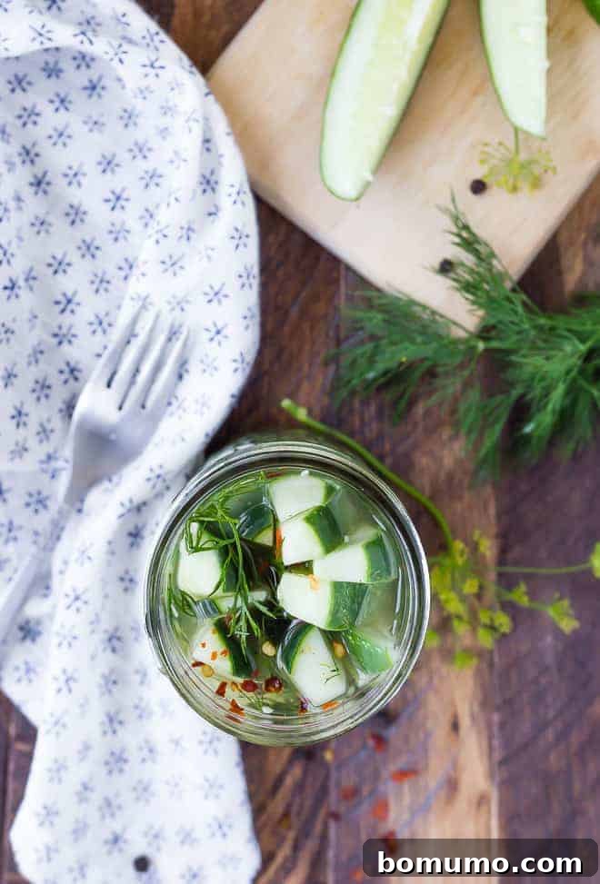 Overhead of pickles in jar, with fork, cloth, fresh dill and sliced cucumbers arranged around.