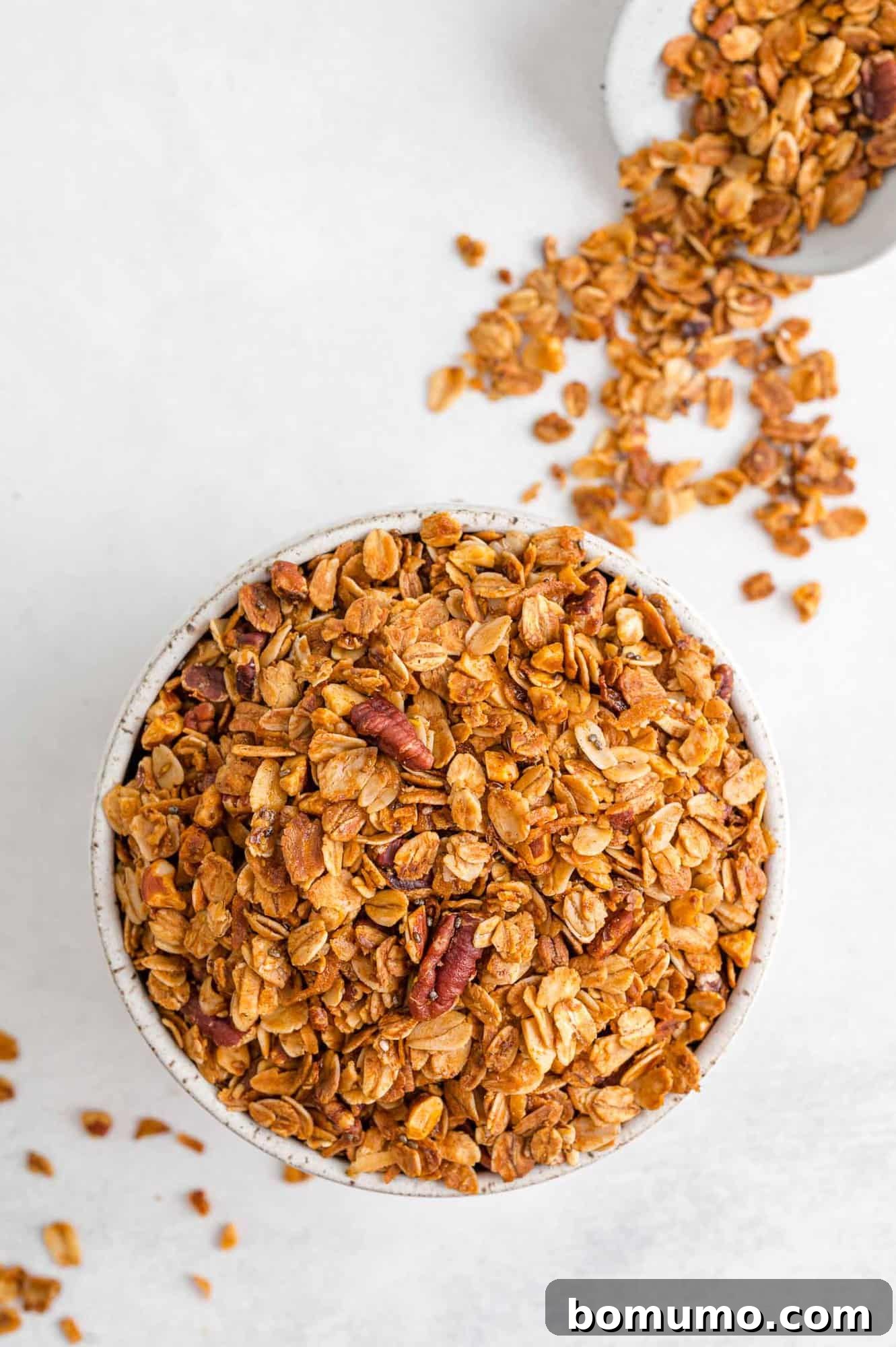 A rustic bowl filled with homemade granola, with more granola scattered on a clean white surface, highlighting its appetizing texture and wholesome appearance.