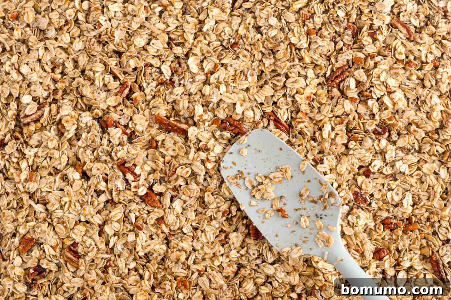 A close-up shot of unbaked granola being meticulously spread into an even, thin layer on a baking pan, demonstrating proper preparation for baking.