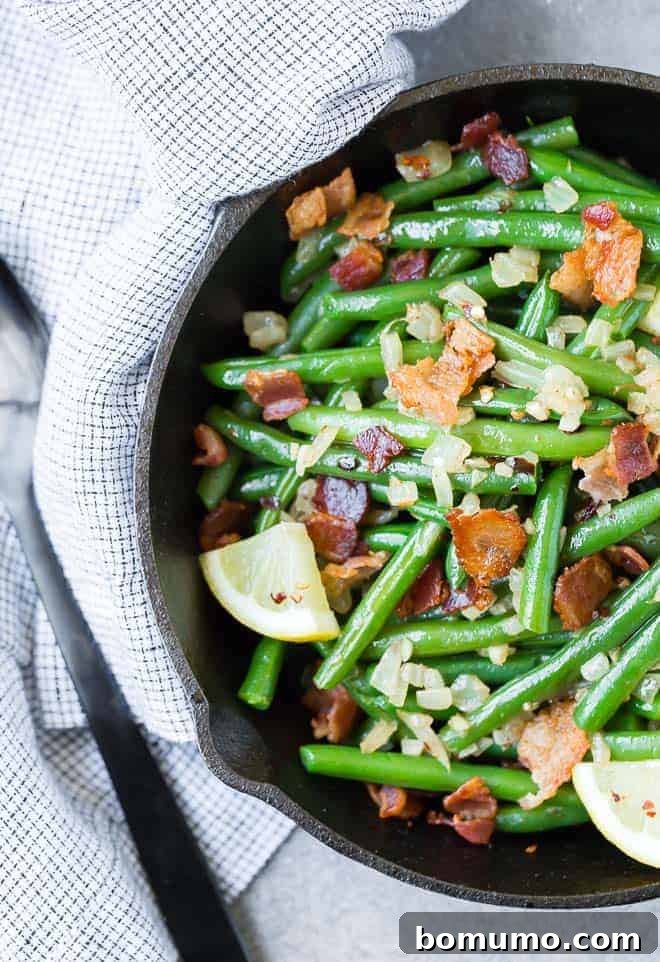 Vibrant green beans in a black cast iron skillet, sprinkled with garlic, onion, and crispy bacon bits. Fresh lemon wedges garnish the dish, adding a burst of color.