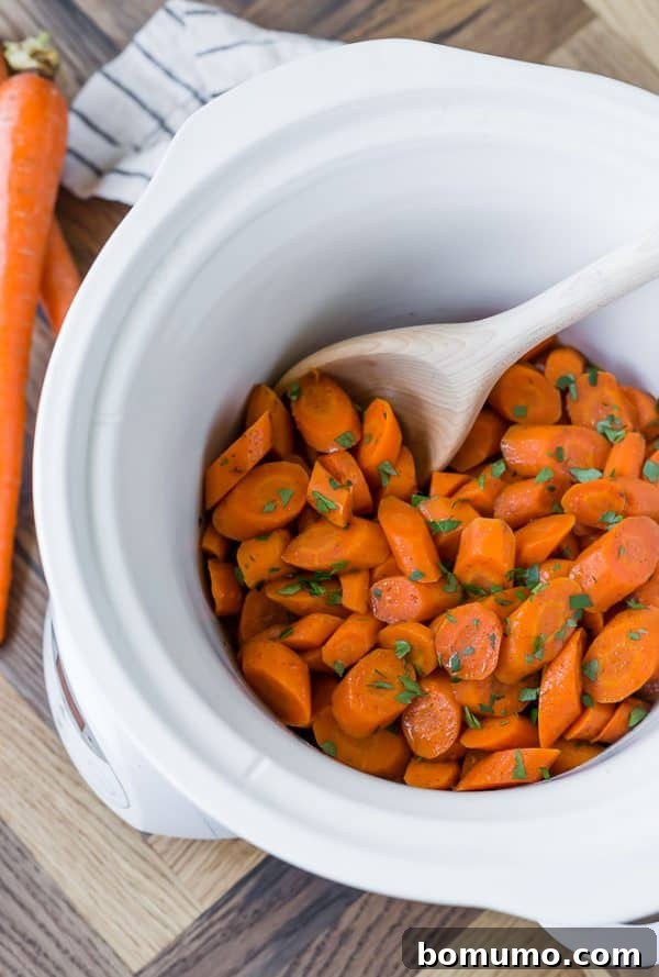 Crockpot glazed carrots in a white slow cooker, with a wooden spoon. Parsley is sprinkled on top.