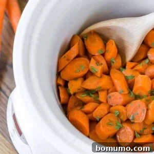 Crockpot glazed carrots in a white slow cooker, with a wooden spoon. Parsley is sprinkled on top.