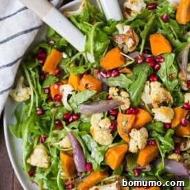 Colorful salad made with arugula, roasted cauliflower, sweet potatoes, shallots, and pomegranate seeds in a large bowl.