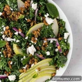 A fresh kale and pear salad in a white bowl, topped with feta cheese, red onions, and savory granola.