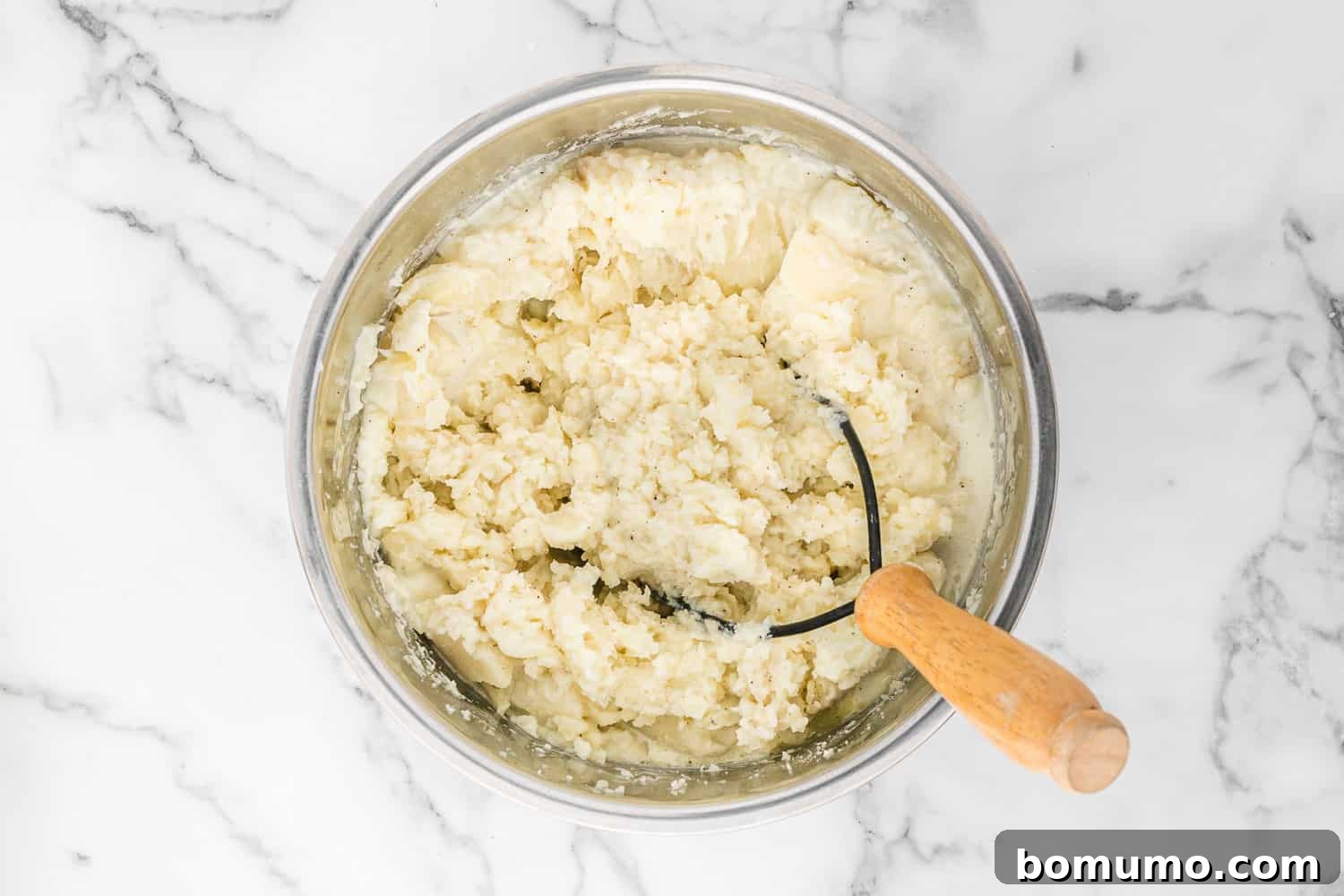 Mashed potatoes with a traditional hand masher, showing a slightly lumpy, rustic texture.