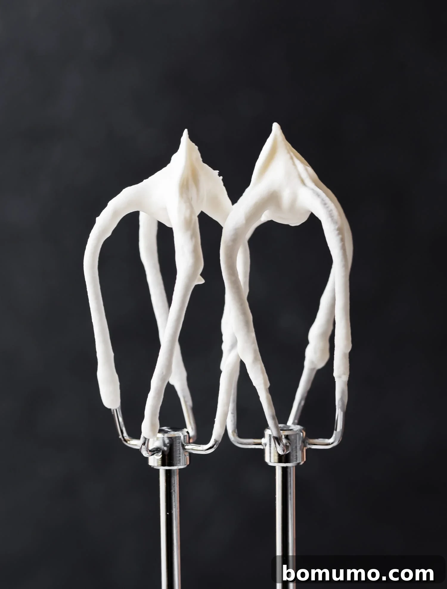 Whipped cream on mixer beaters, against black background, illustrating ideal texture.