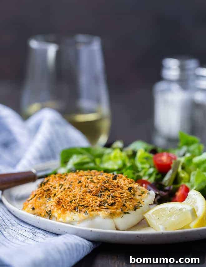 Two beautifully baked parmesan cod fillets on a white plate, garnished with fresh herbs, with a blurred glass of white wine in the background.