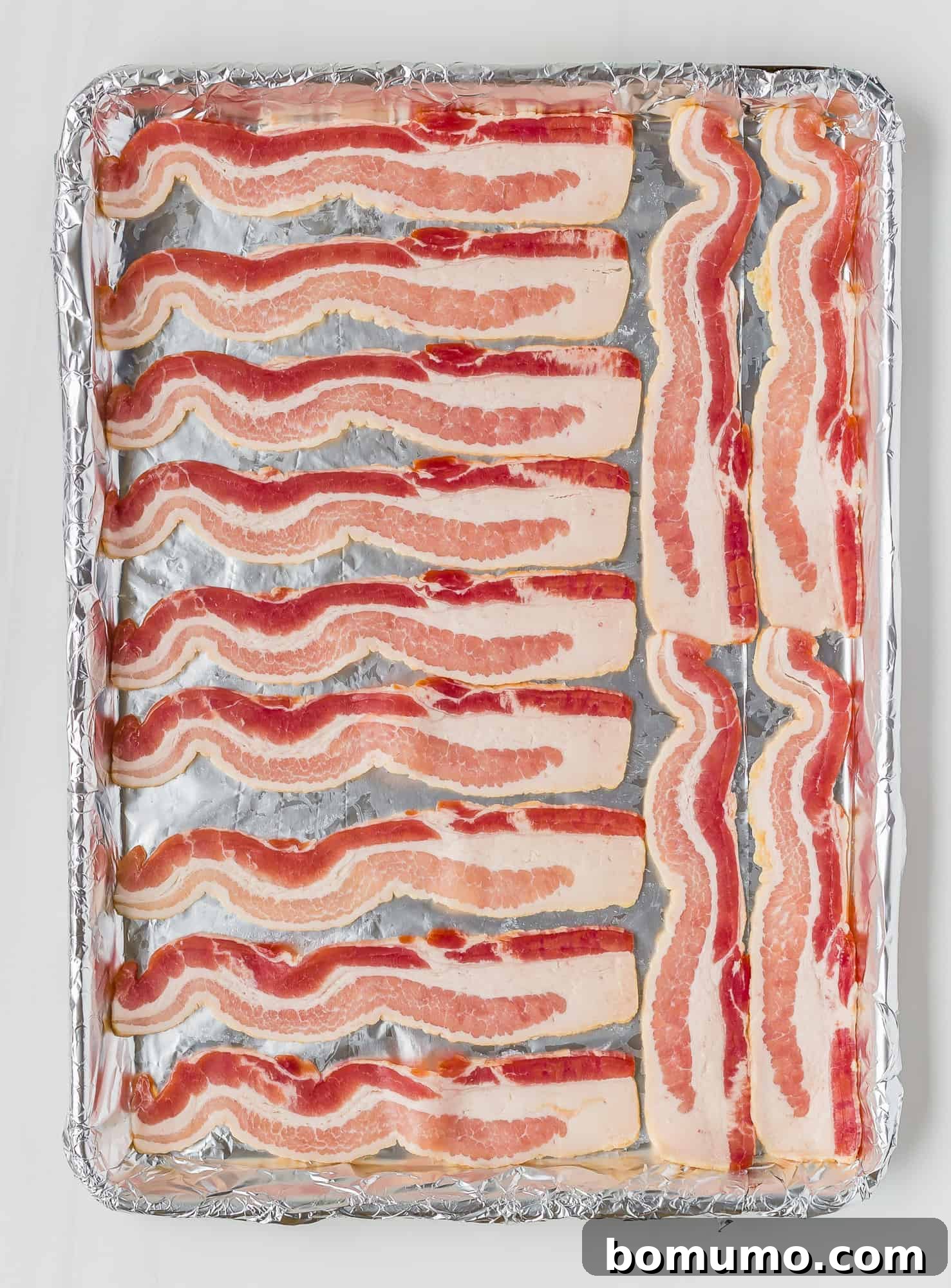 Uncooked bacon strips laid out in a single layer on a foil-lined baking sheet, ready for the oven. Each strip is distinct, promising even cooking.