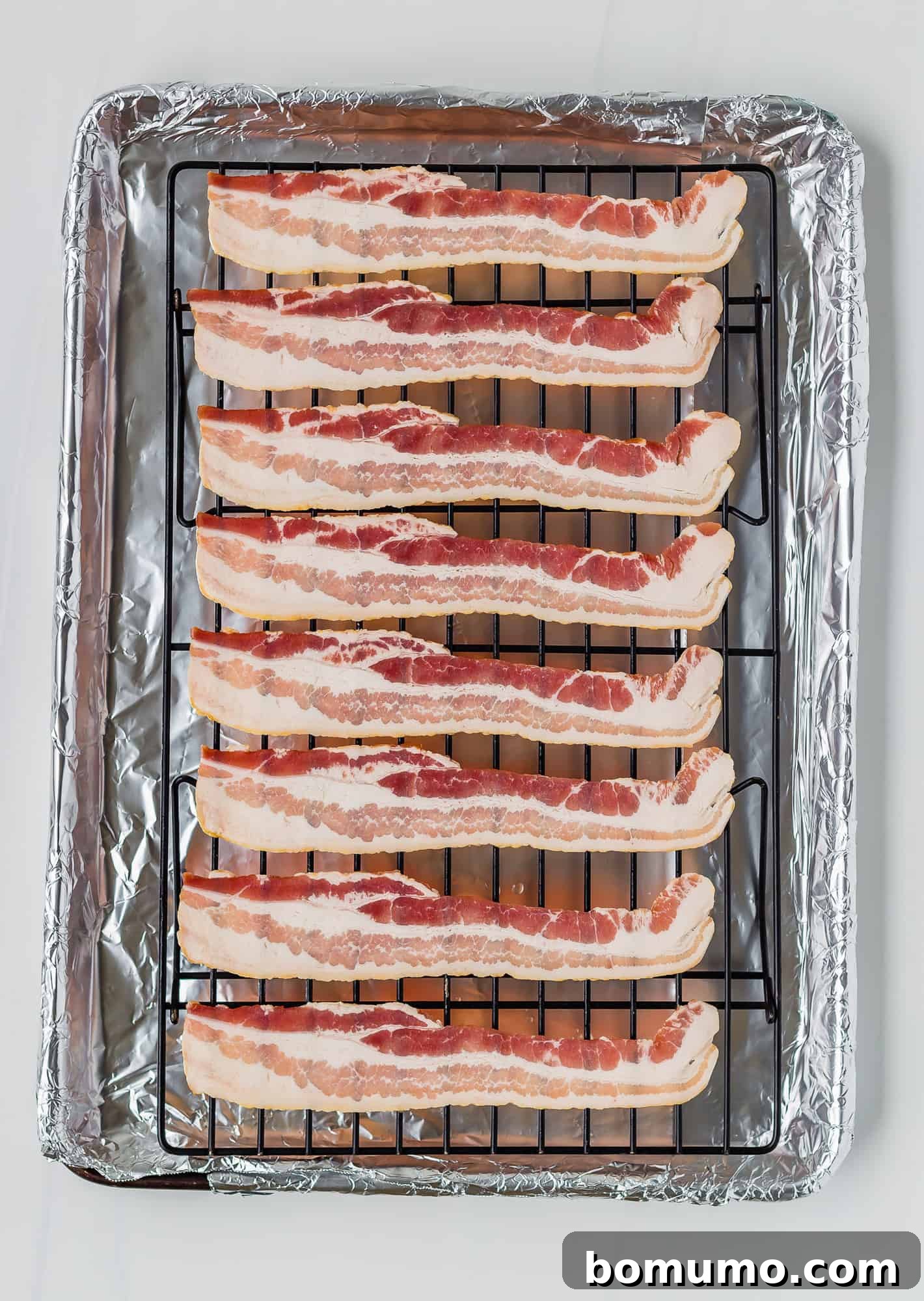 Uncooked bacon strips arranged on a wire rack placed over a foil-lined baking sheet, ready for baking. The spacing ensures even air circulation.
