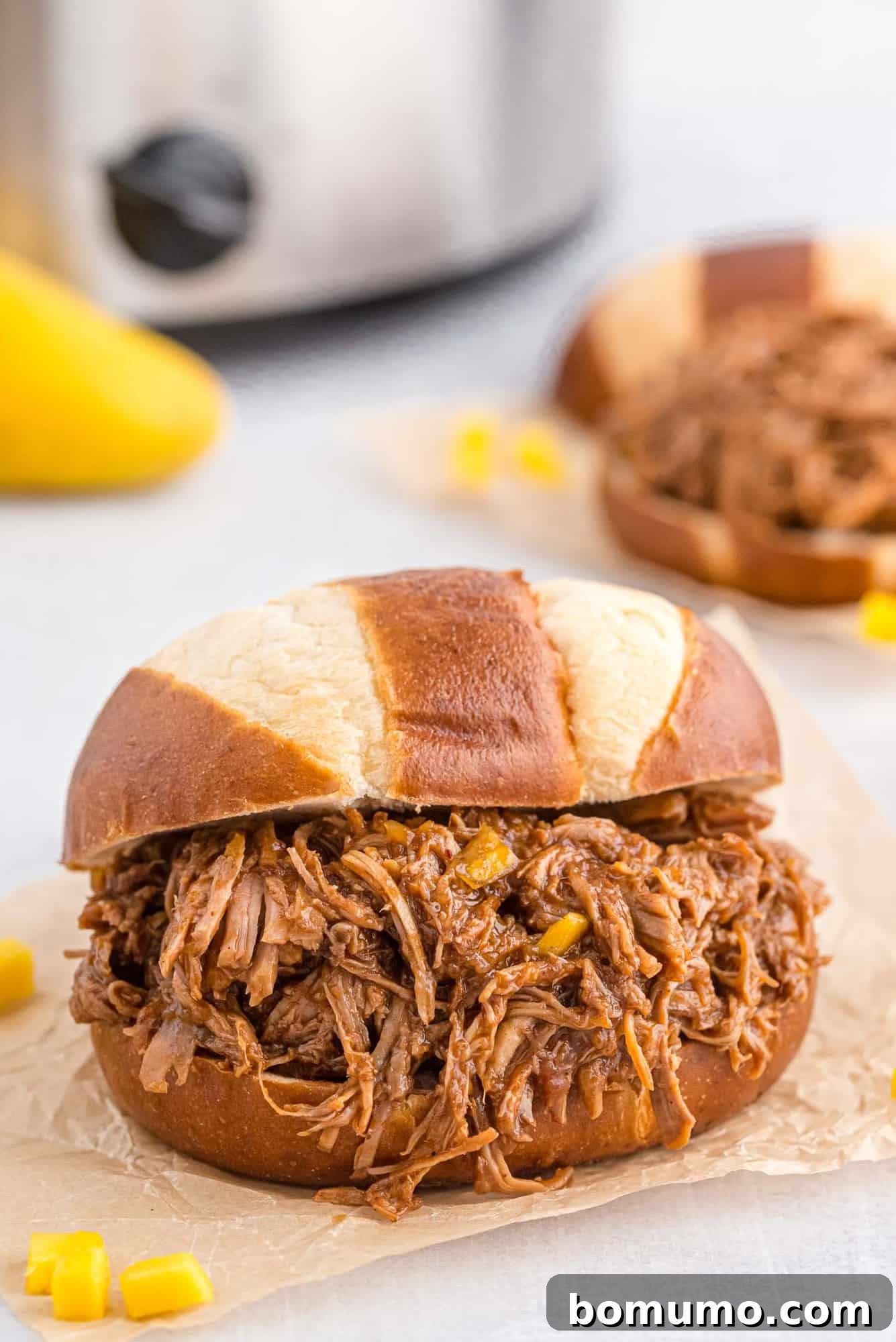Pretzel bun piled high with pulled pork, a slow cooker pictured in the background.