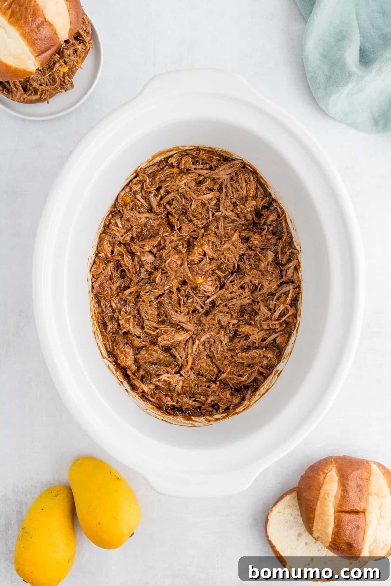 Pork in a slow cooker surrounded by mangos and pretzel rolls.