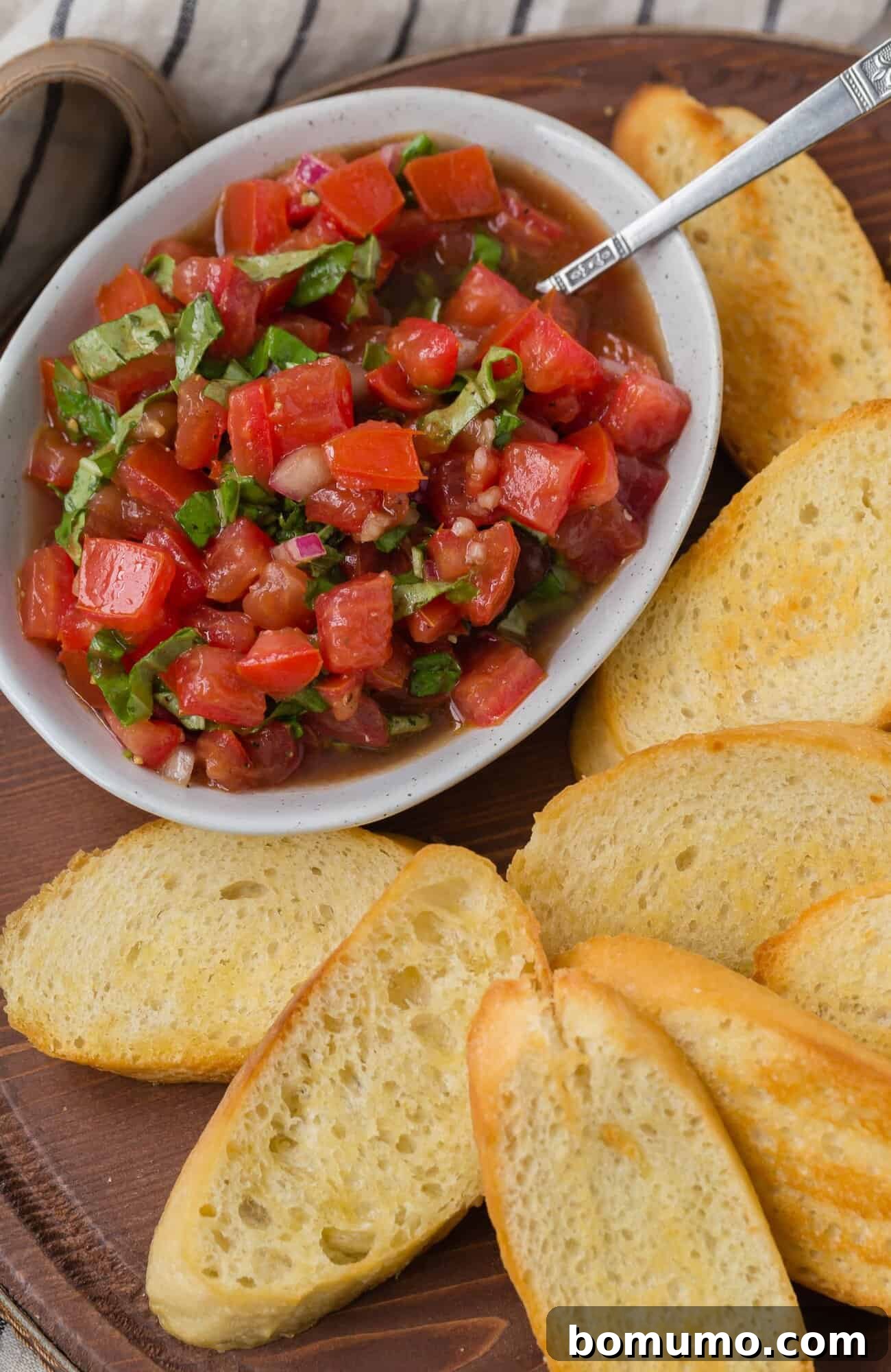 A bowl of vibrant tomato bruschetta topping, featuring freshly chopped tomatoes, fragrant basil, and a hint of red onion, artfully arranged beside several golden slices of toasted baguette on a rustic wooden board.