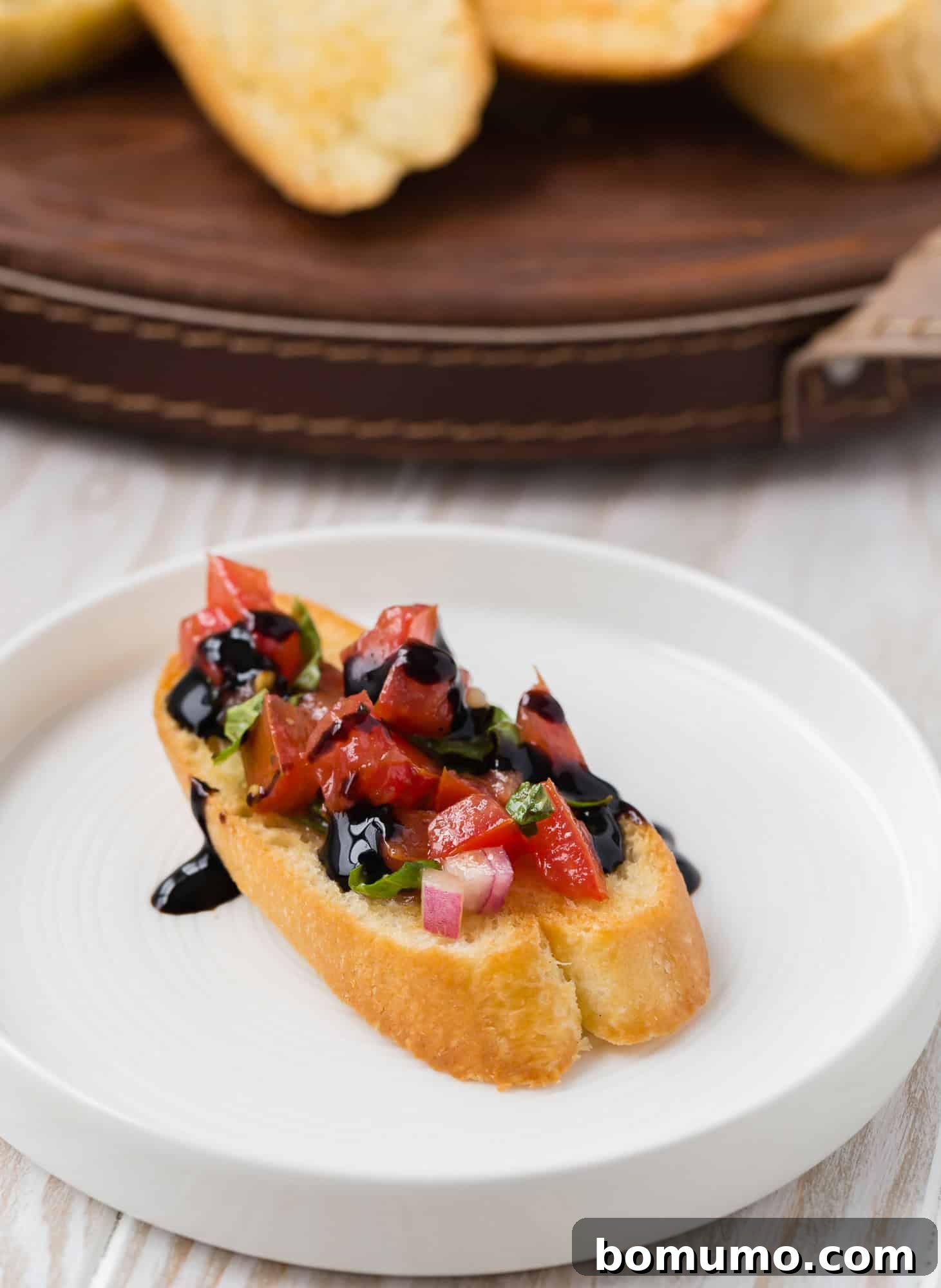 A perfectly toasted crostini topped with a vibrant mix of diced tomatoes, red onion, and fresh basil, elegantly finished with a drizzle of rich balsamic glaze, presented on a pristine white plate.