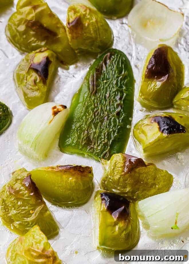 Image of perfectly roasted tomatillos, onions, and jalapeños on a foil-lined sheet pan, ready for blending.