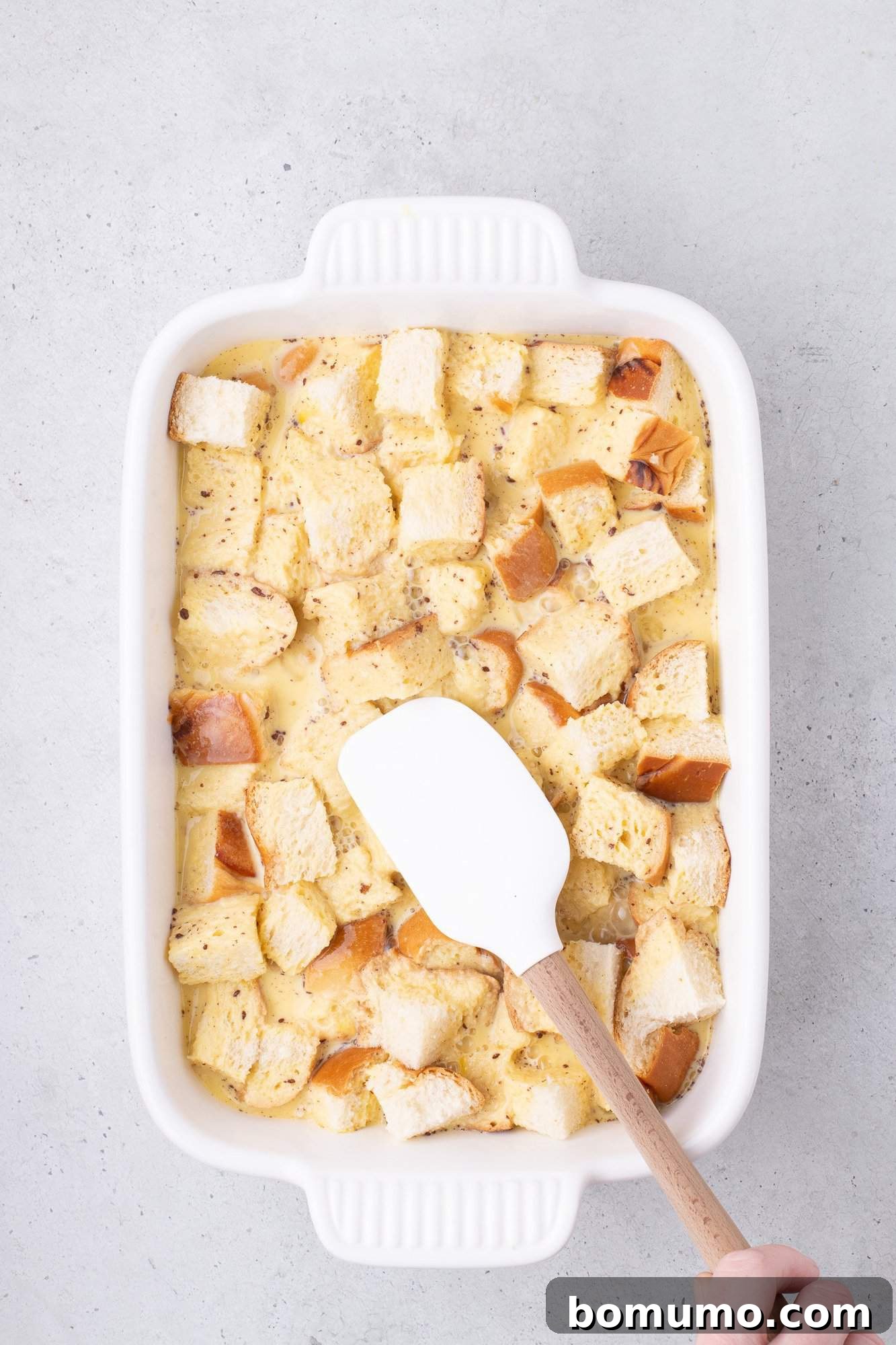 A white baking dish contains cubed bread soaked in a creamy mixture. A white spatula is in contact with the bread.