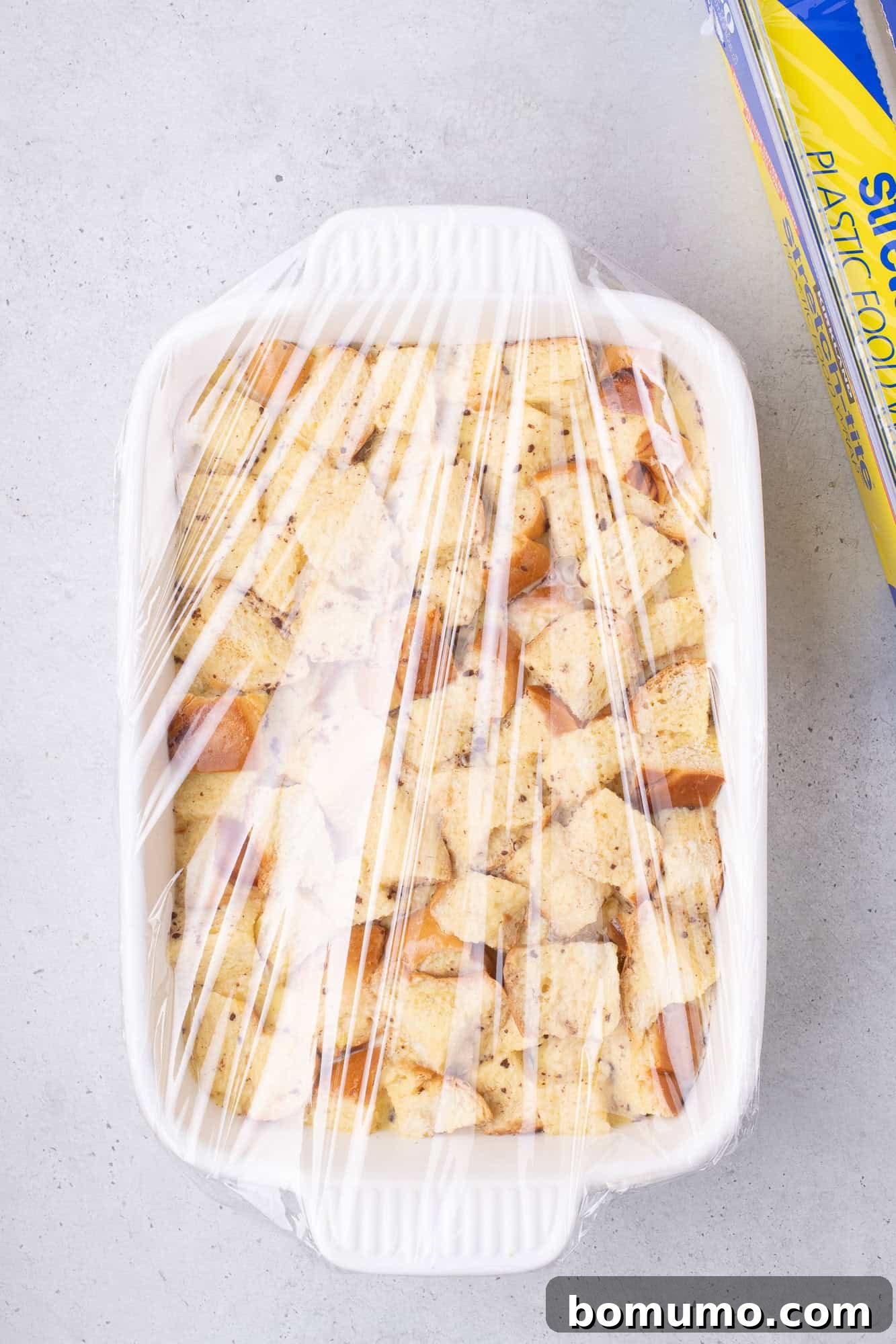 A white casserole dish filled with cubed bread is wrapped in plastic wrap. A box of plastic wrap is nearby on a gray surface.