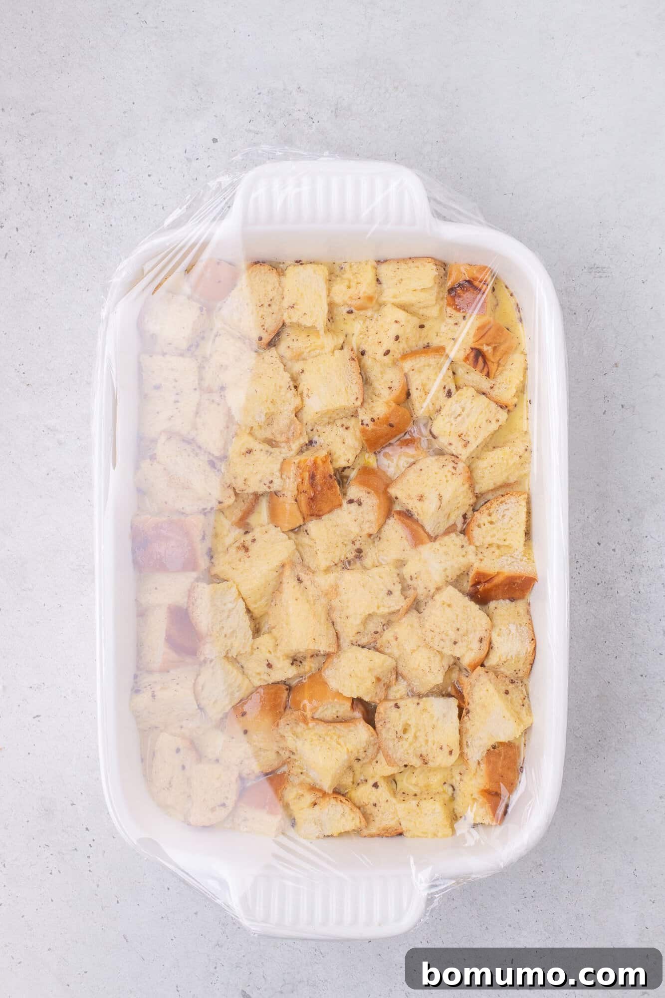 A white baking dish filled with cubed bread covered with plastic wrap, ready for baking.
