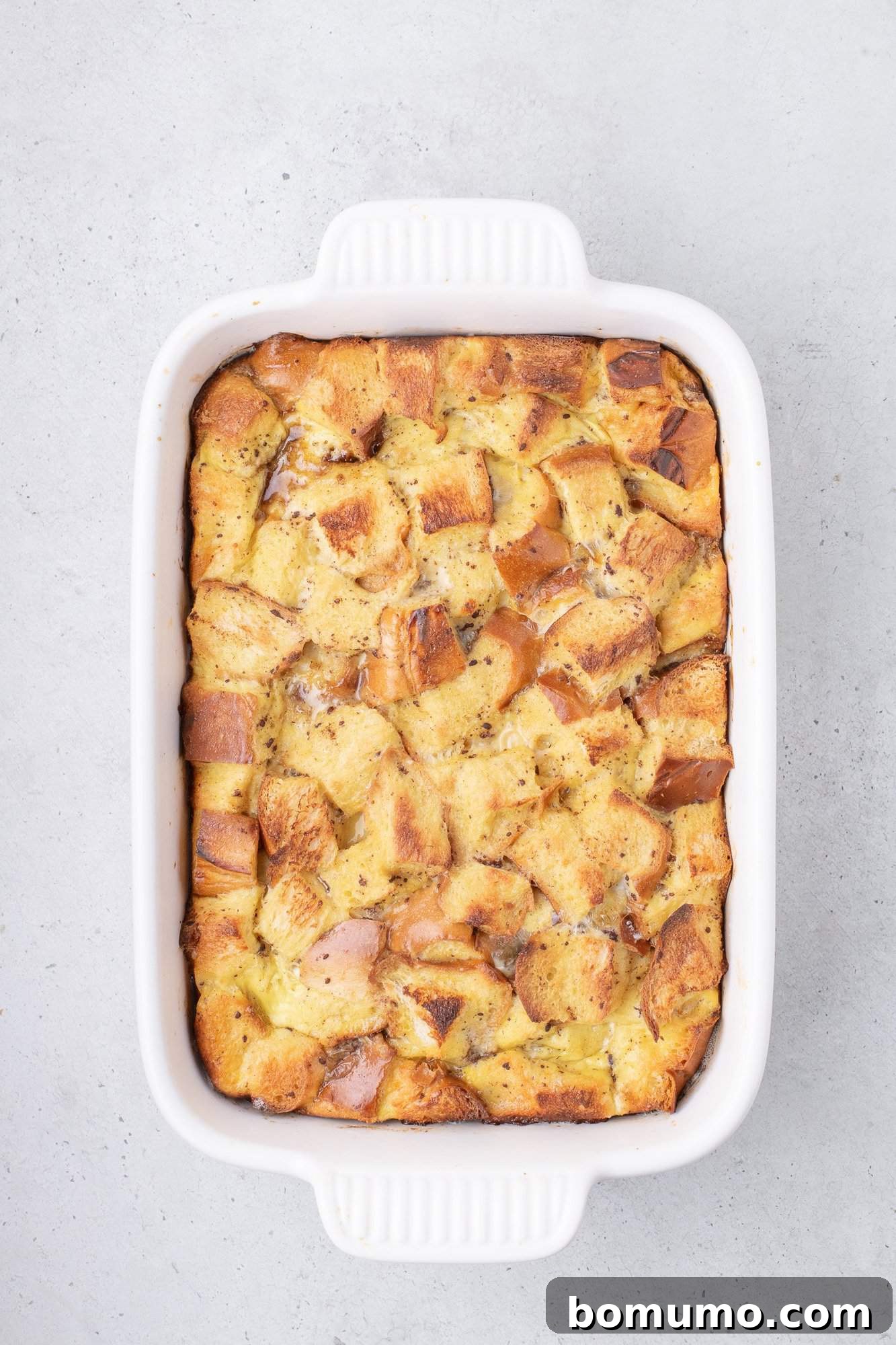 A baked french toast in a white rectangular dish, featuring golden brown bread cubes in a creamy mixture, on a light gray surface.