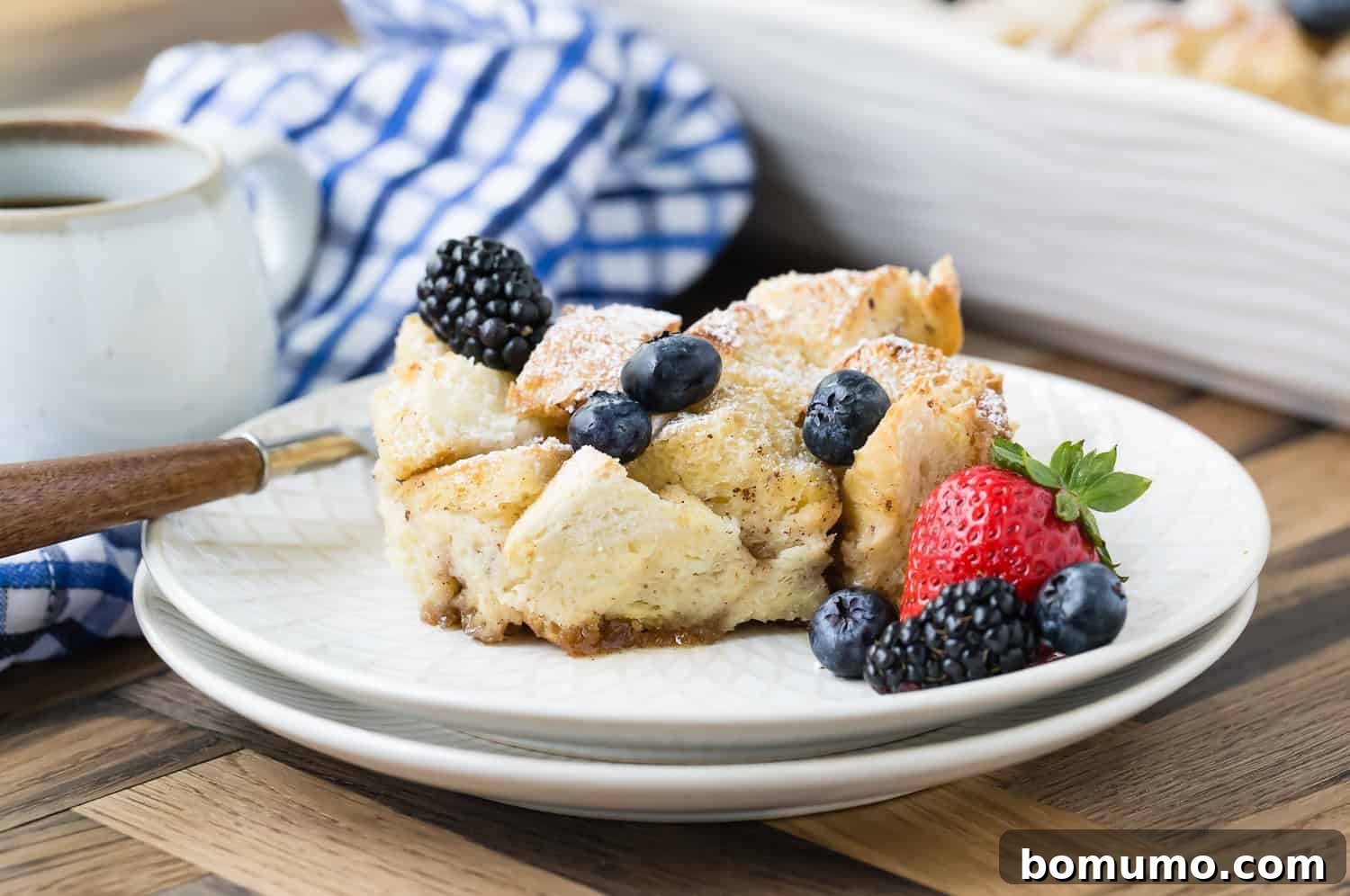 A serving of bread pudding topped with powdered sugar, blueberries, blackberries, and a strawberry, on a white plate with a fork. 