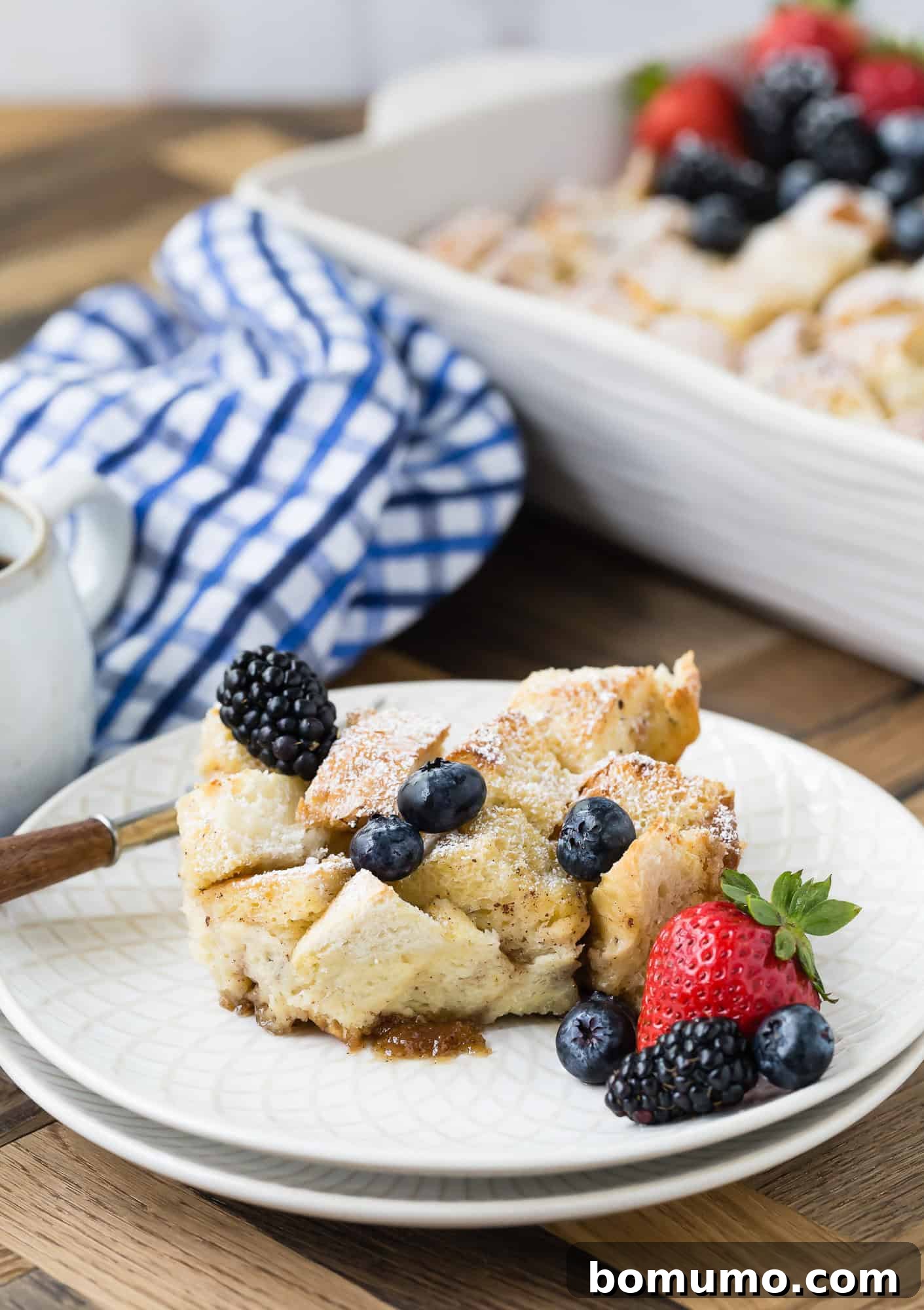 A slice of bread pudding on a plate, topped with blackberries and blueberries. A casserole dish and a blue checkered cloth are in the background.