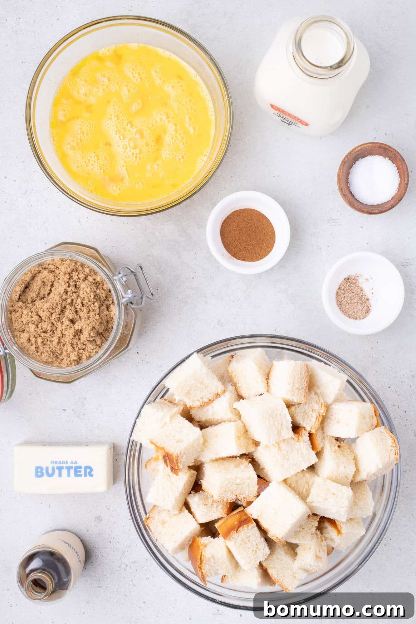 Ingredients for recipe are laid out: cubed bread, beaten eggs, milk, brown sugar, butter, cinnamon, and salt, arranged on a light-colored surface.