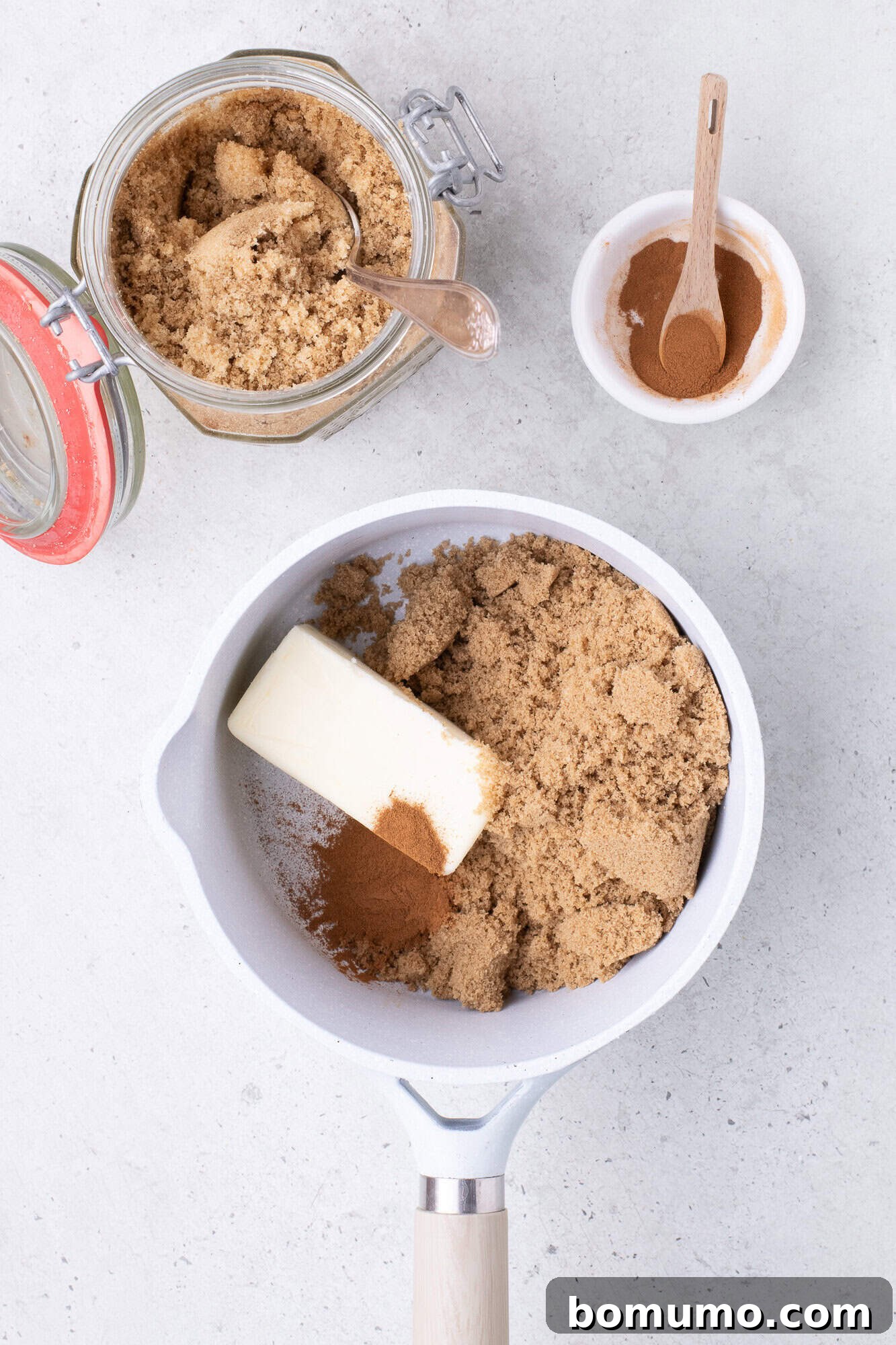 Butter, brown sugar, and cinnamon in a white saucepan, with extra sugar in a jar and cinnamon in a small bowl nearby on a light surface.