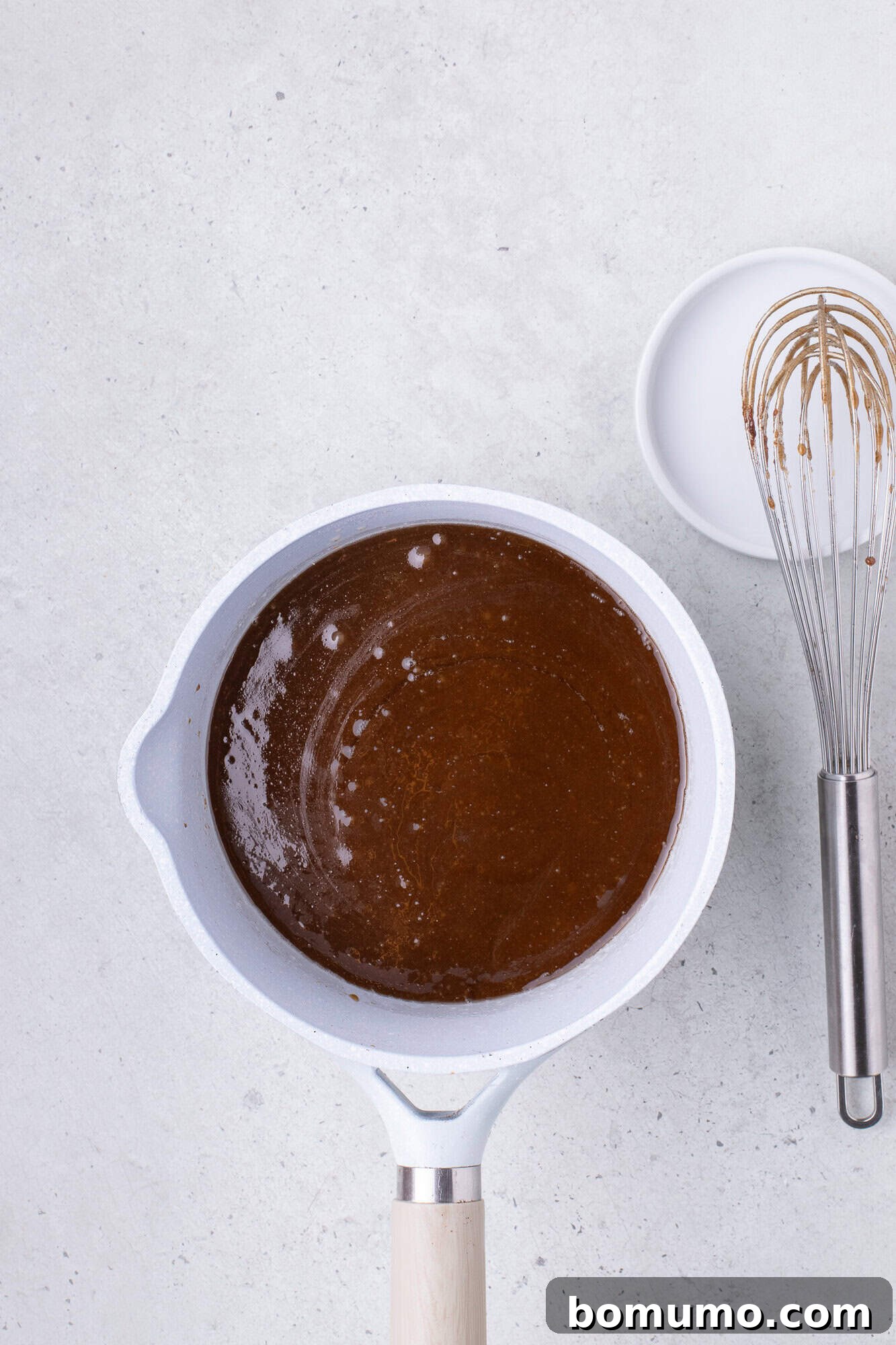 A white pot containing chocolate sauce sits on a light countertop, next to a whisk and an empty white bowl.