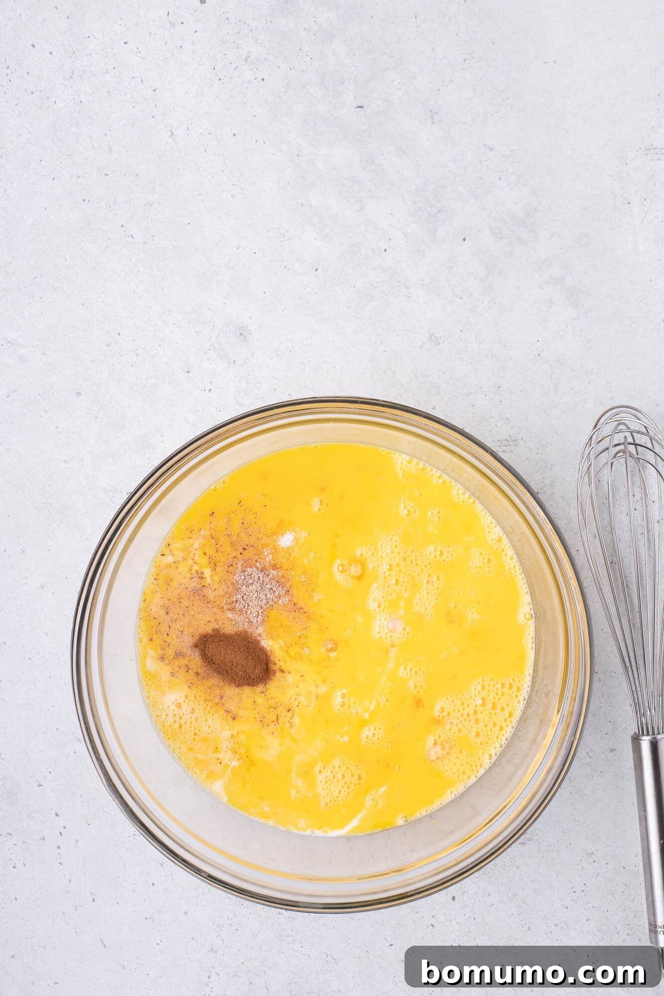 A glass bowl with beaten eggs and spices next to a metal whisk on a gray countertop.