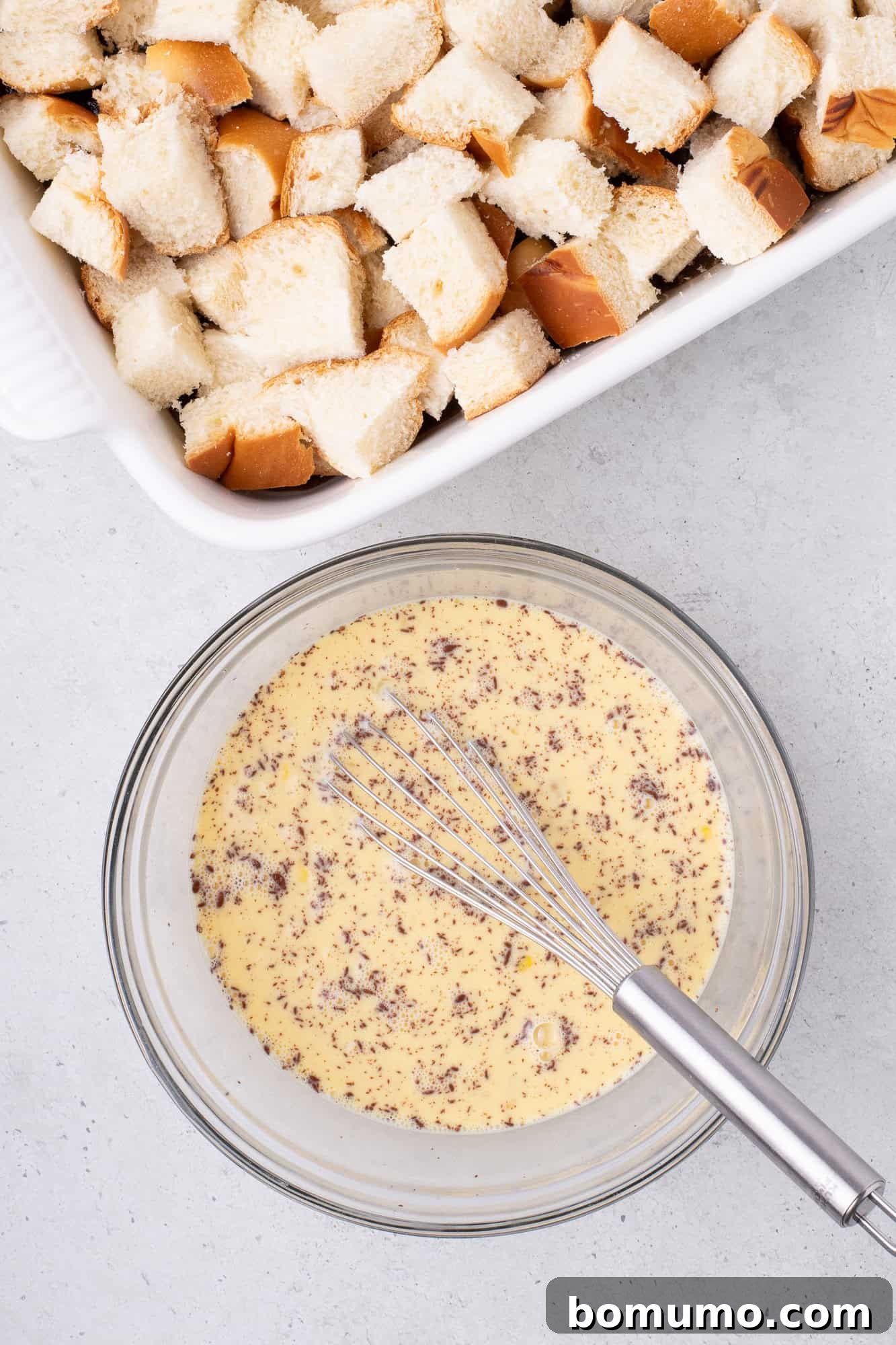 Cubed bread in a white baking dish; a glass bowl with a whisk and a speckled egg mixture beside it on a gray surface.