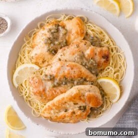 A bowl of cooked angel hair pasta topped with seared chicken piccata and a lemon-caper sauce, garnished with lemon slices.
