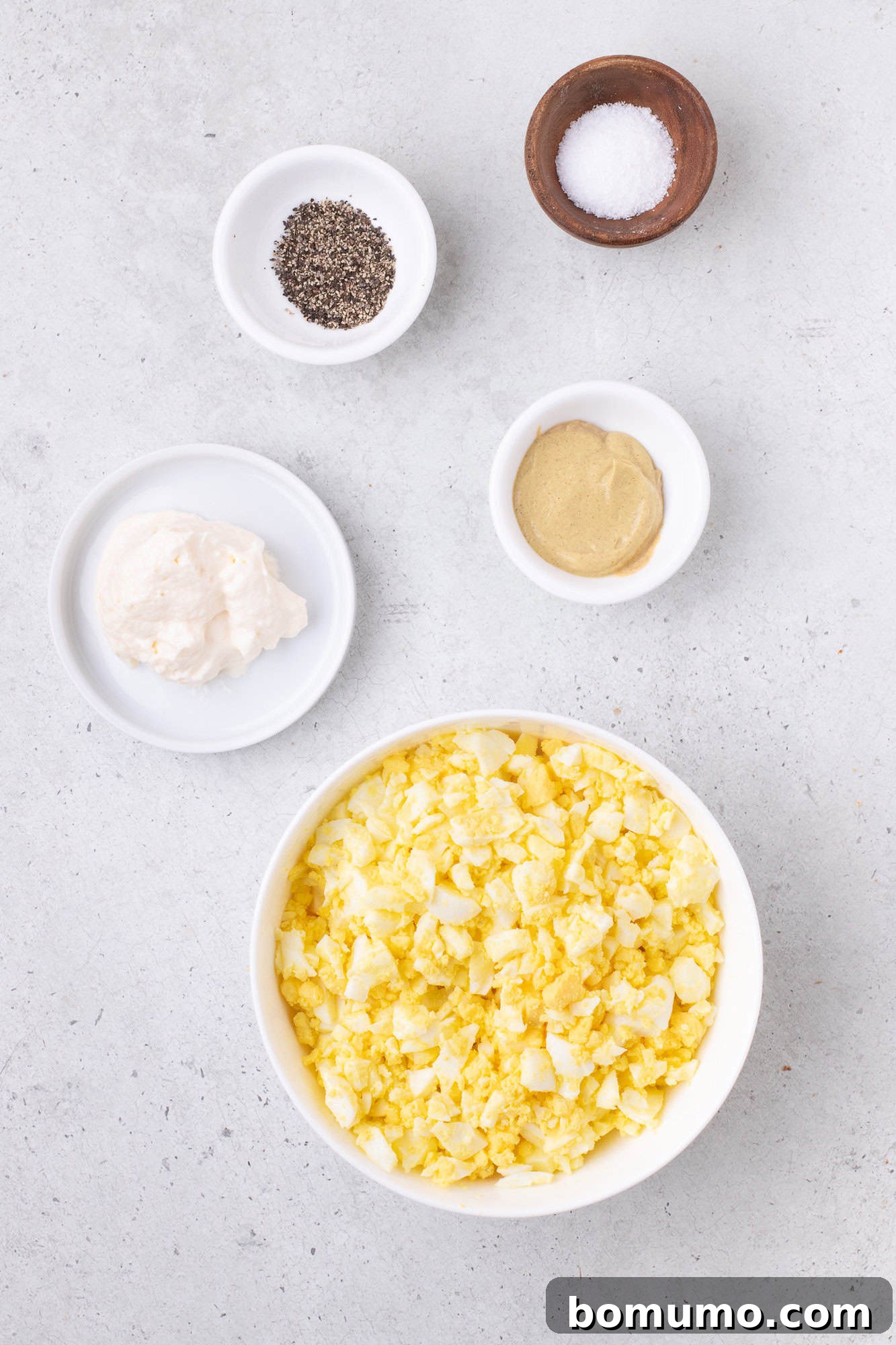 Healthy Egg Salad Makeover 4 Chopped hard-boiled eggs in a bowl, surrounded by small dishes of creamy mayonnaise, tangy mustard, and a sprinkle of salt and pepper, ready to be mixed.