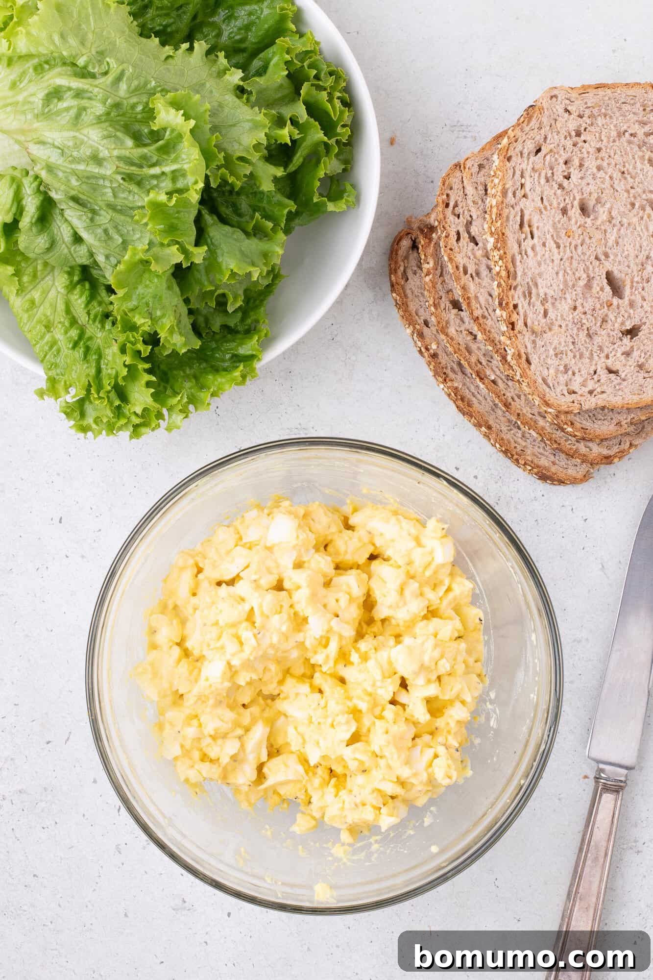 Healthy Egg Salad Makeover 6 A bowl of creamy egg salad ready to be served, alongside a plate of crisp lettuce leaves, wholesome brown bread slices, and a knife on a white culinary surface.